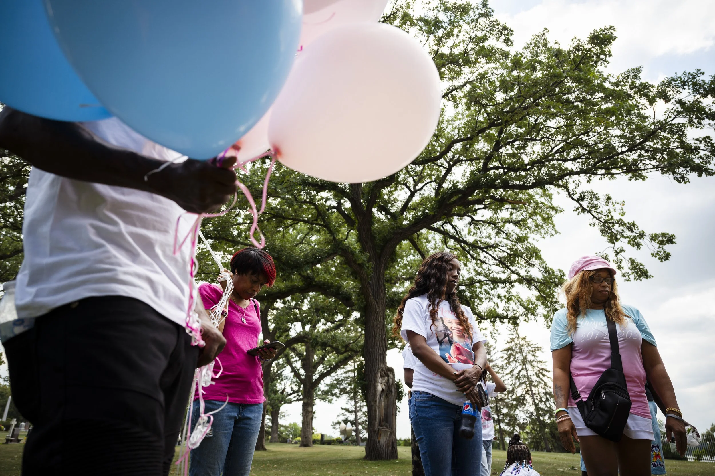 A group of people standing outdoors in a park, holding helium balloons in pink and blue colors. The background features tall trees and a cloudy sky.