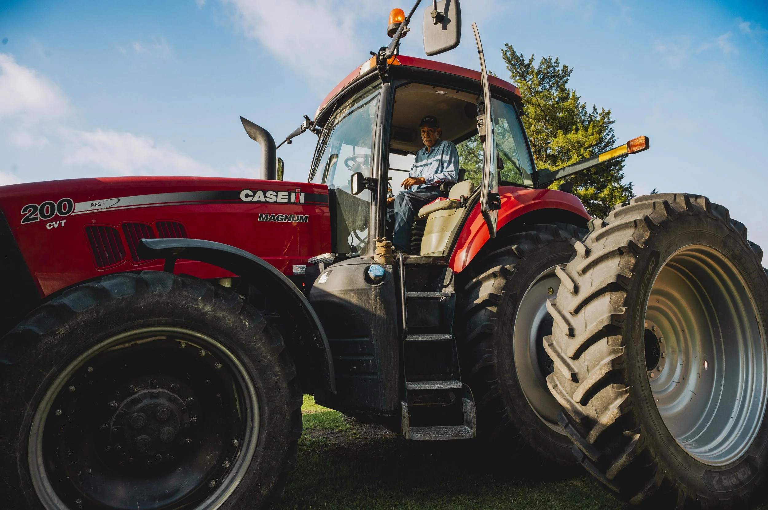 A man operating a large red CASE IH Magnum 200 tractor outdoors with trees and a blue sky in the background.