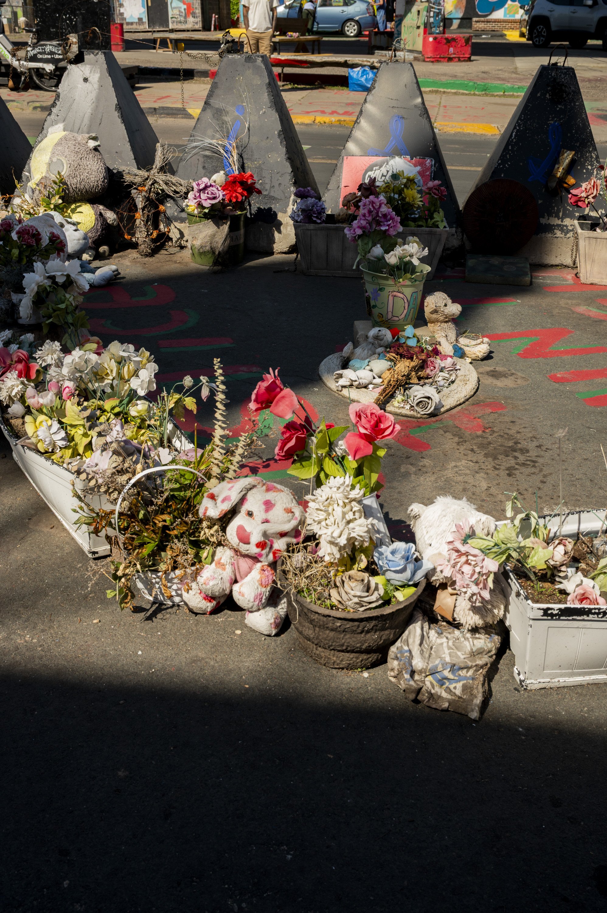 Flowers, stuffed toys, candles, and memorial items placed on the ground in front of concrete barriers, indicating a memorial or tribute site.