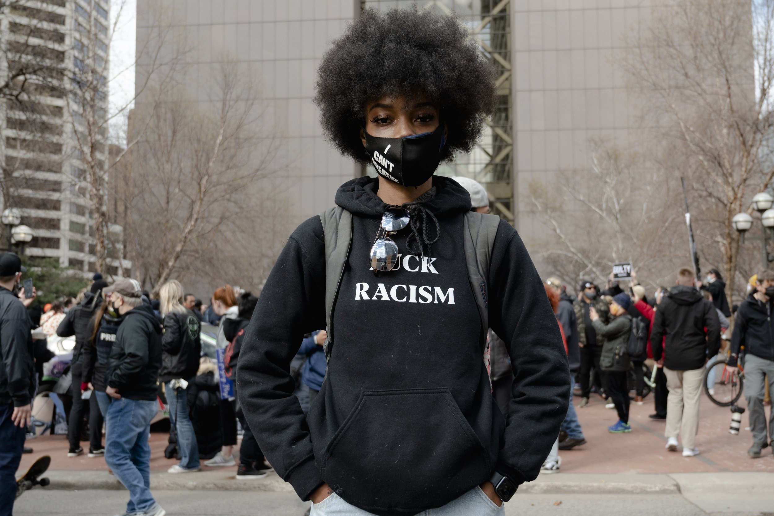 A woman wearing a black mask, black hoodie, and carrying a backpack at a protest, with a crowd of protesters and trees in the background.