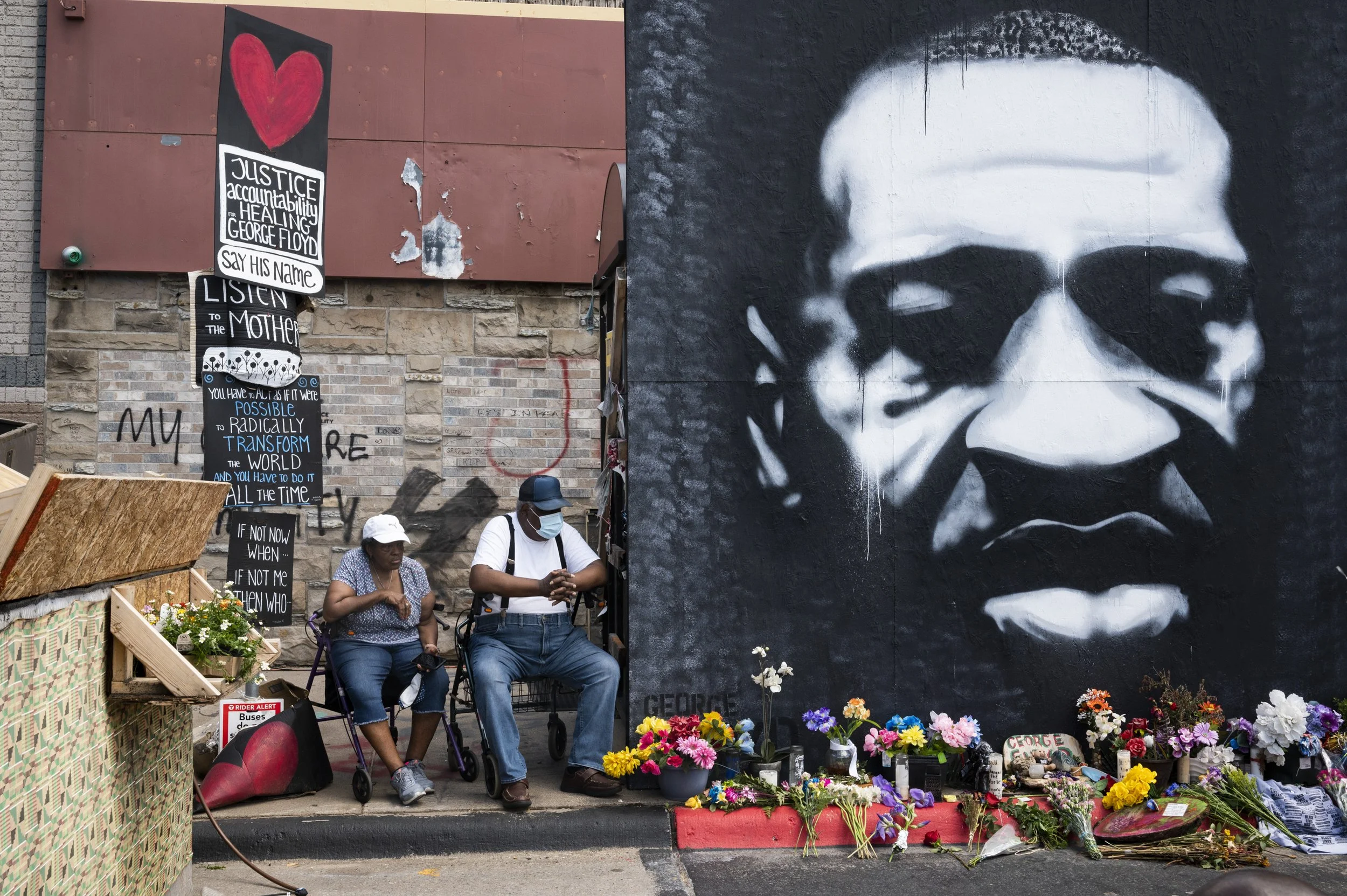 Street memorial with flowers and artwork honoring George Floyd, featuring a large black and white portrait of George Floyd and posters with messages about justice and racial transformation, two people sitting nearby with masks.