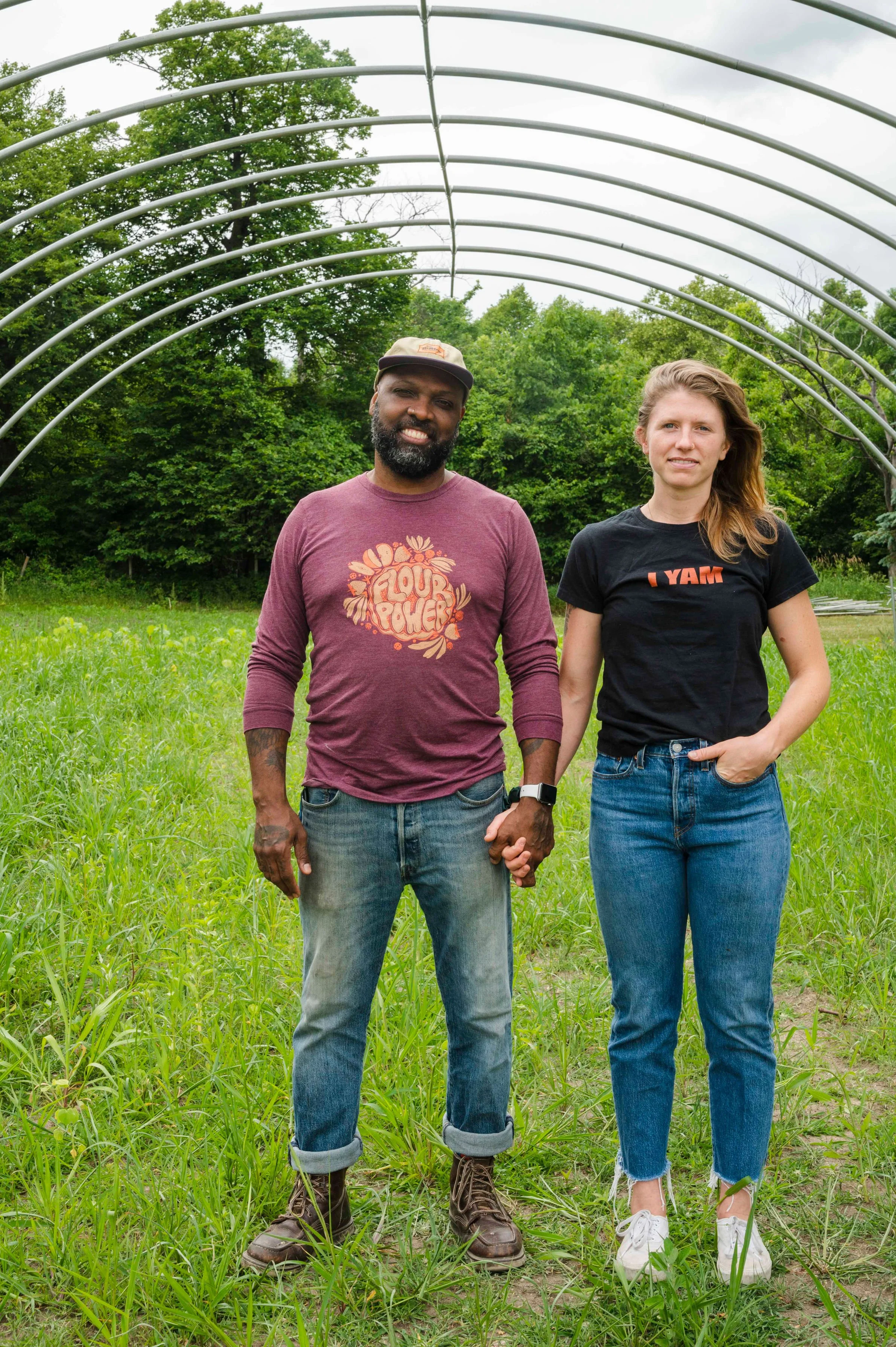 A man and woman holding hands in an outdoor green field with a metal arch structure above them and trees in the background.