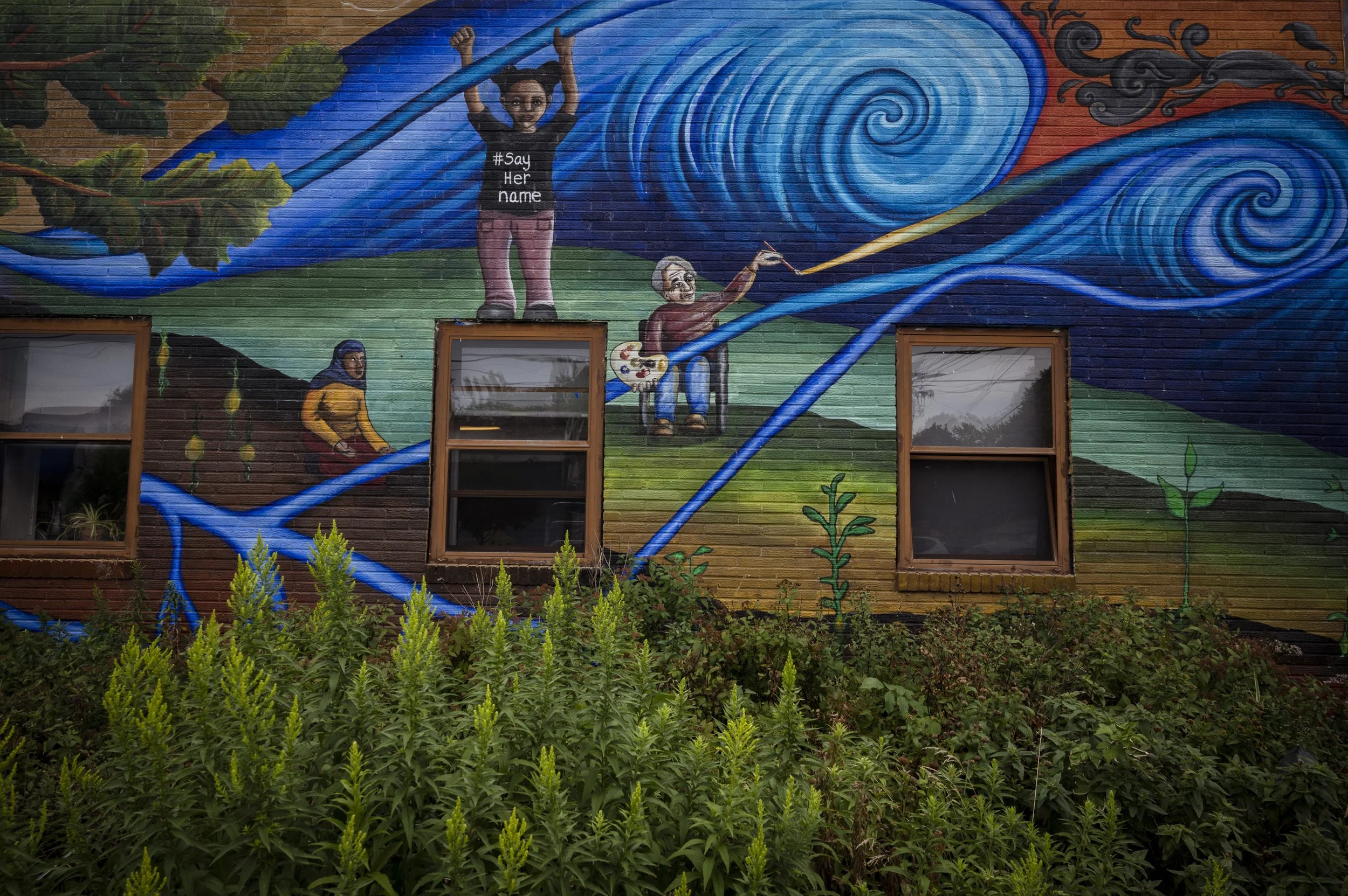 A colorful mural on a brick wall depicting a woman with two buns in her hair holding a long stick, with symbols resembling wind or water swirls, a person with a palette painting, and plants in the foreground.