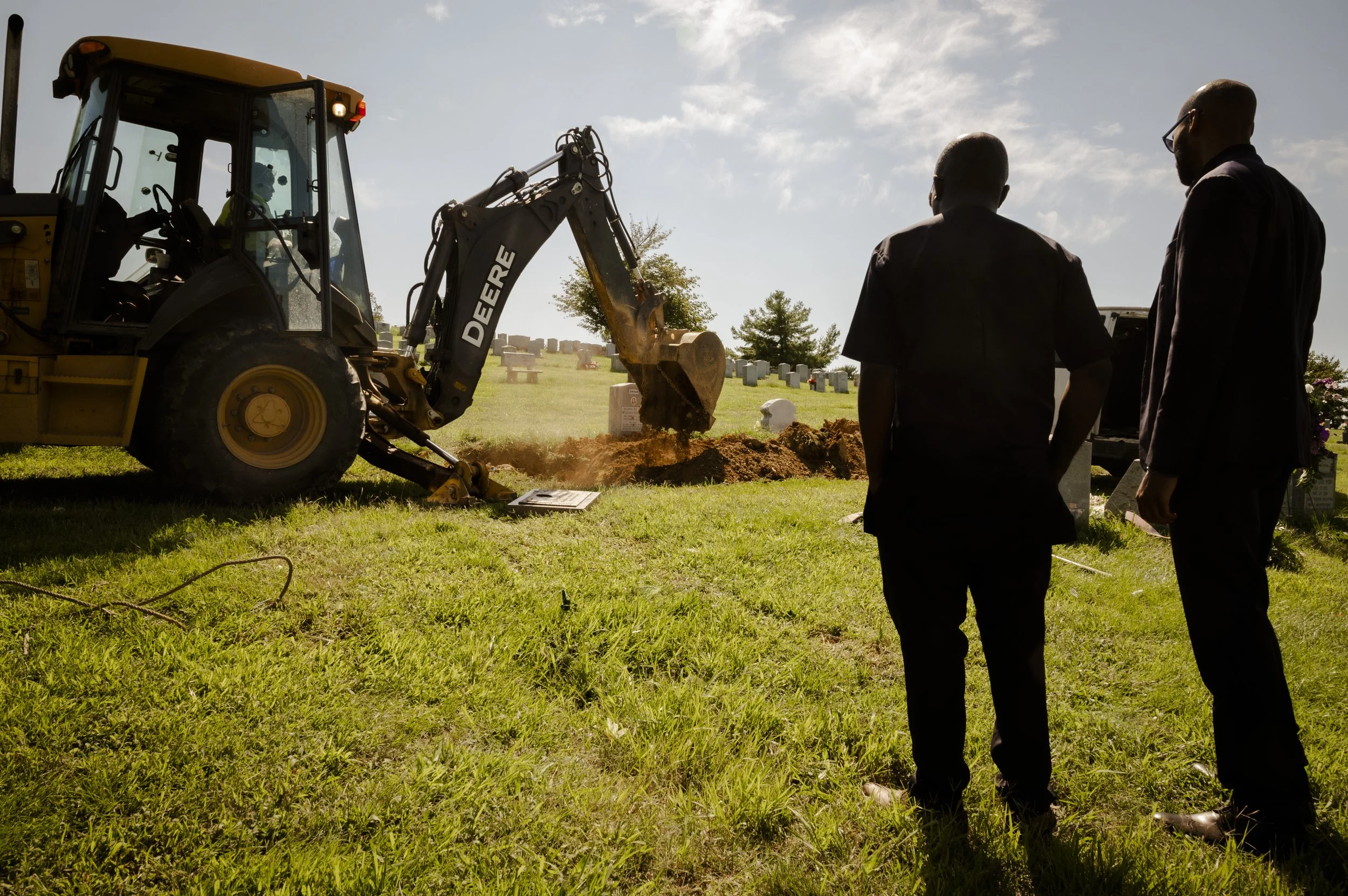 Two men in black business suits stand in a grassy cemetery, observing a construction site where a yellow bulldozer is digging a grave, with headstones and trees in the background.