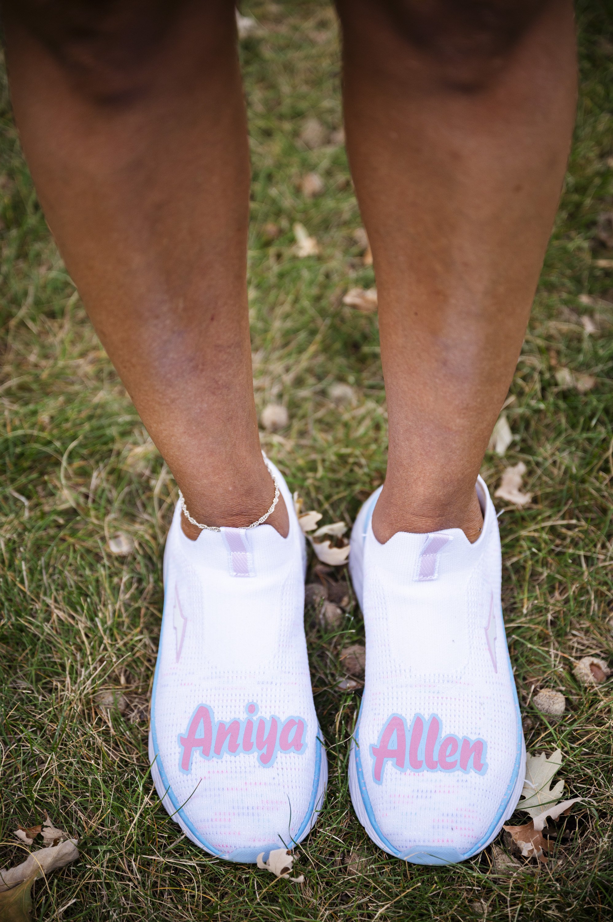 Pair of white slip-on shoes with pink lettering displaying names 'Aniya' and 'Allen' on the ground with grass and fallen leaves.