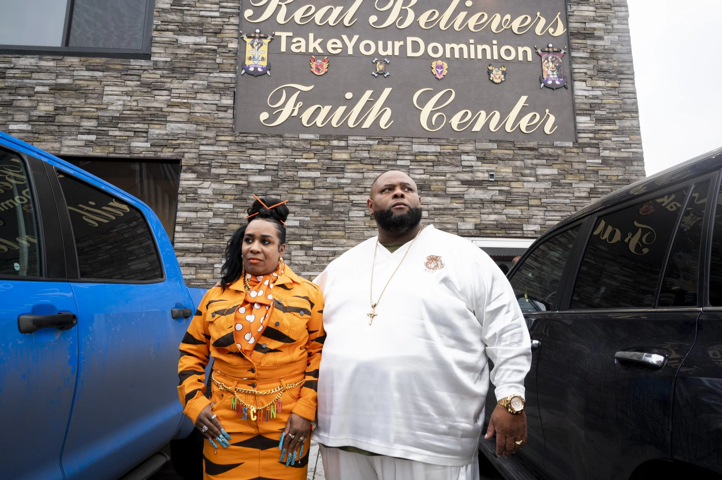A man and woman standing outside a church named Faith Center with a sign that reads 'Real Believers Take Your Dominion.' The woman wears an orange and black tiger-striped outfit with a polka-dot scarf and statement jewelry, and has her hair styled with accessories. The man is dressed in a white outfit with a gold watch and necklace, and has a beard. They stand between blue and black vehicles.