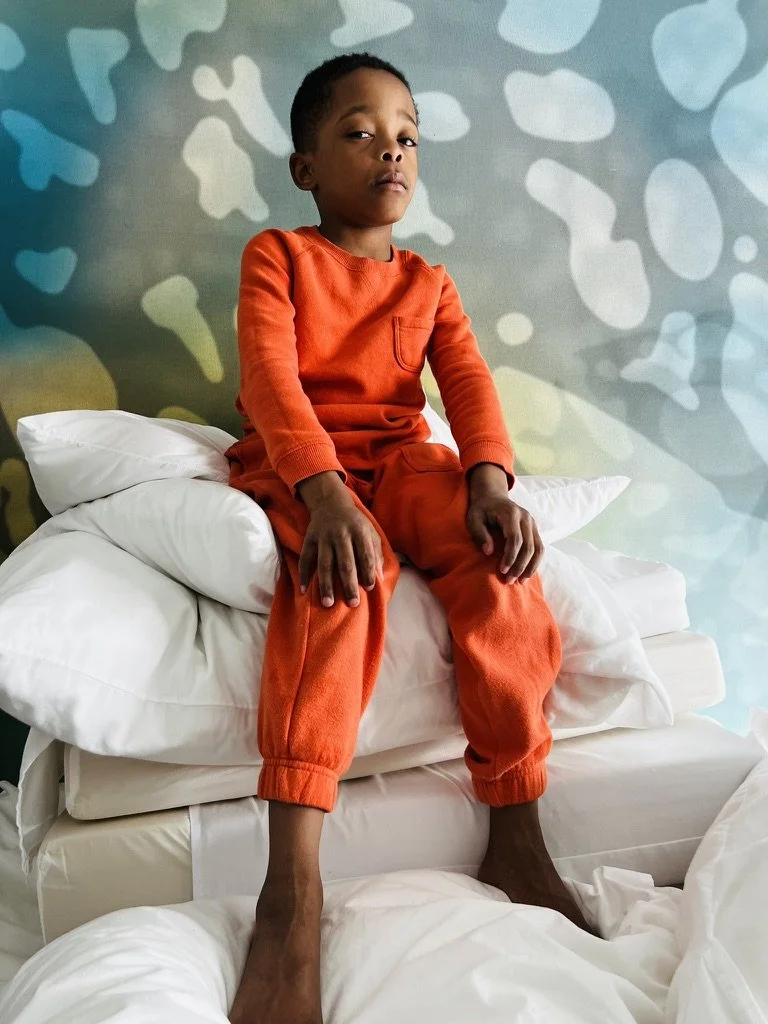A young boy sits on a stack of white pillows on a bed, wearing an orange long-sleeve shirt and orange pants, with a serious expression, against a colorful abstract background.