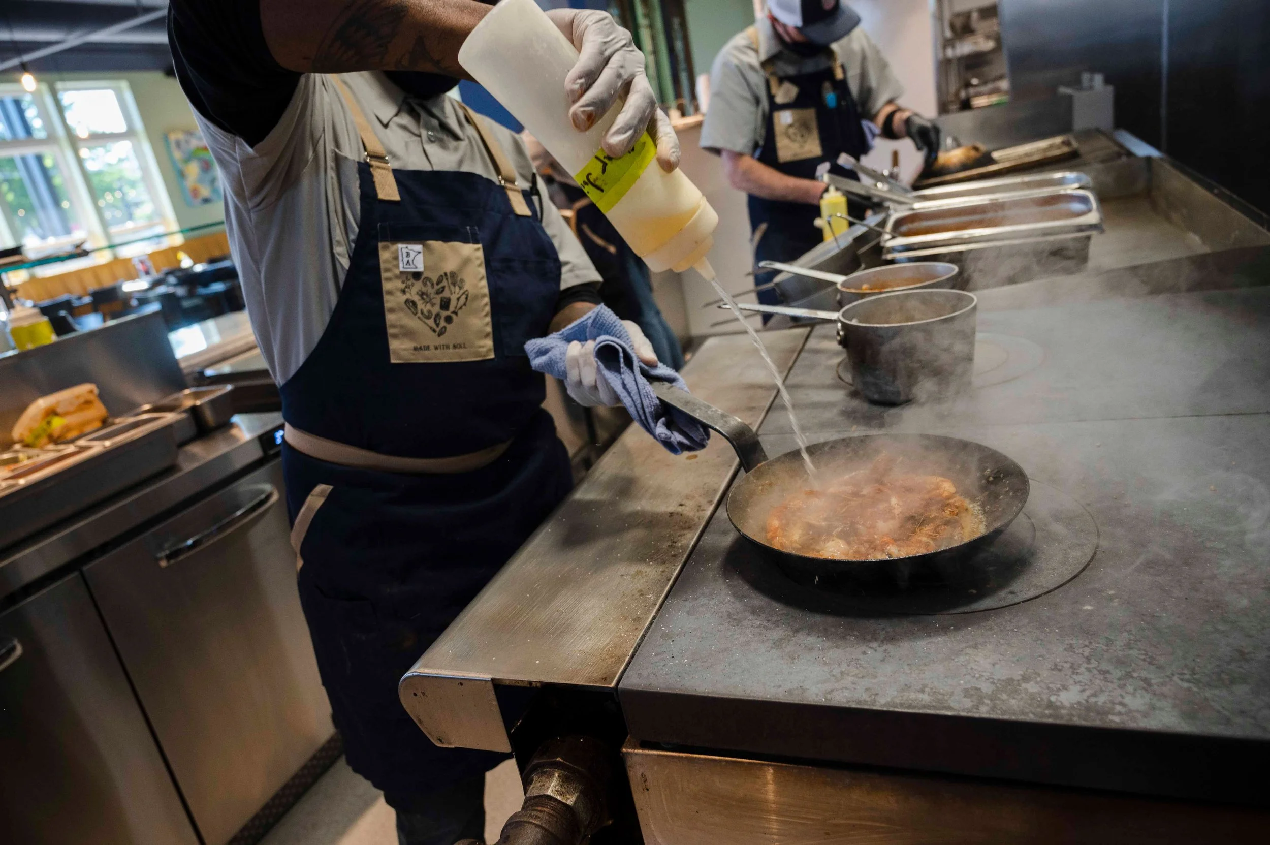 Chef cooking on a stovetop, pouring sauce into a skillet with steam rising, in a busy restaurant kitchen.
