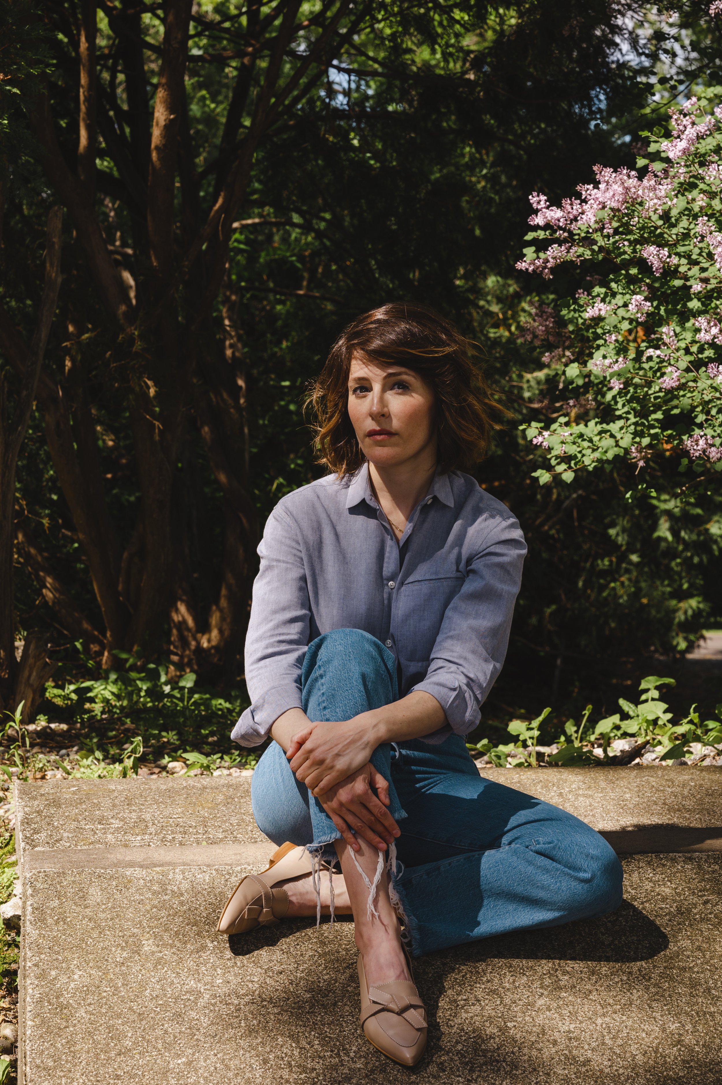 White woman with wavy brown hair, wearing a gray button-up shirt and blue jeans, sitting on a concrete step in a garden with trees and pink flowering bushes.