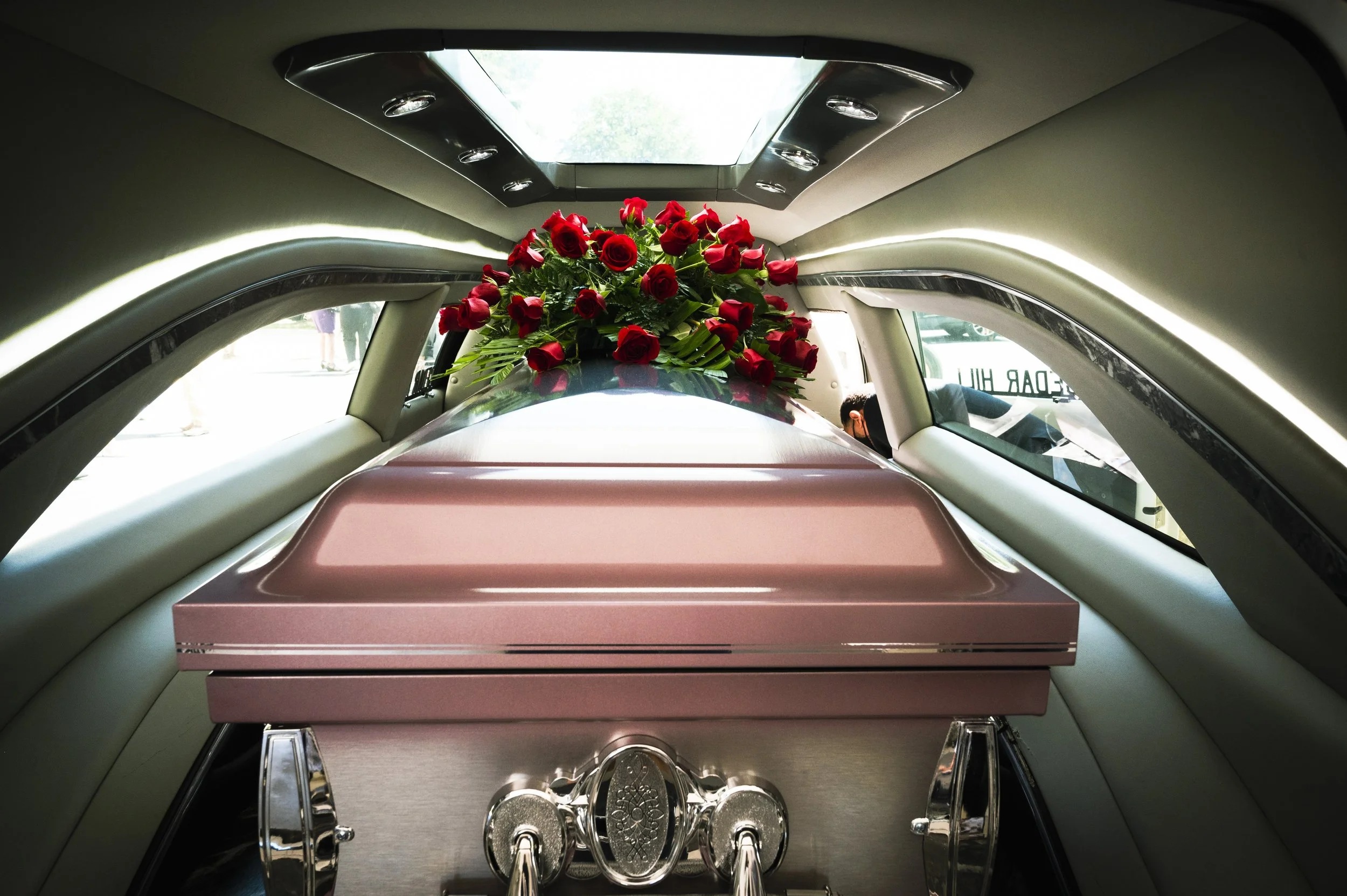 A pink coffin inside a hearse, decorated with a bouquet of red roses and green leaves, viewed from the rear.