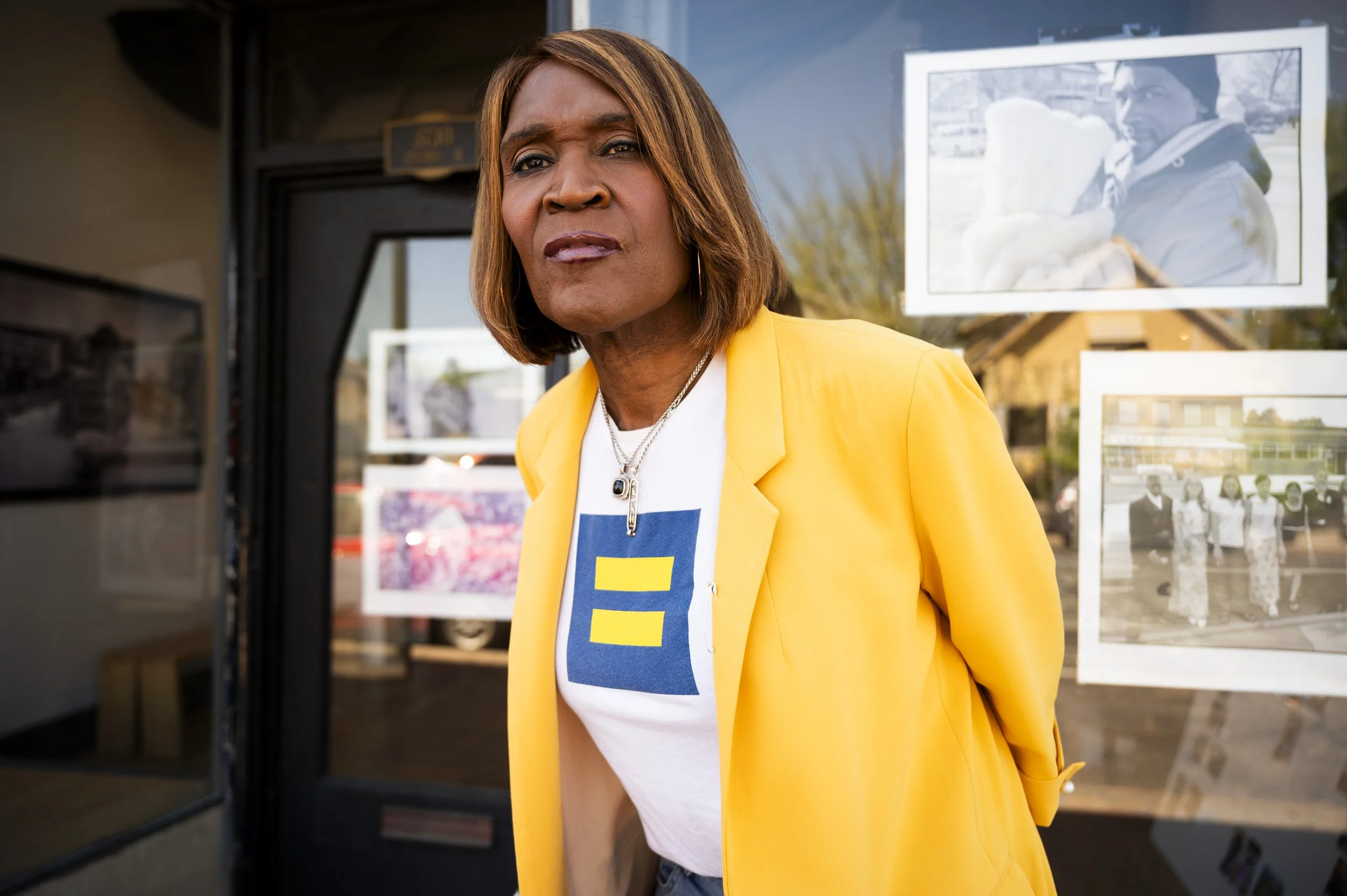 Woman with shoulder-length hair wearing a yellow blazer and a white T-shirt with a colorful design, standing outside a storefront with photos of people displayed in the window.