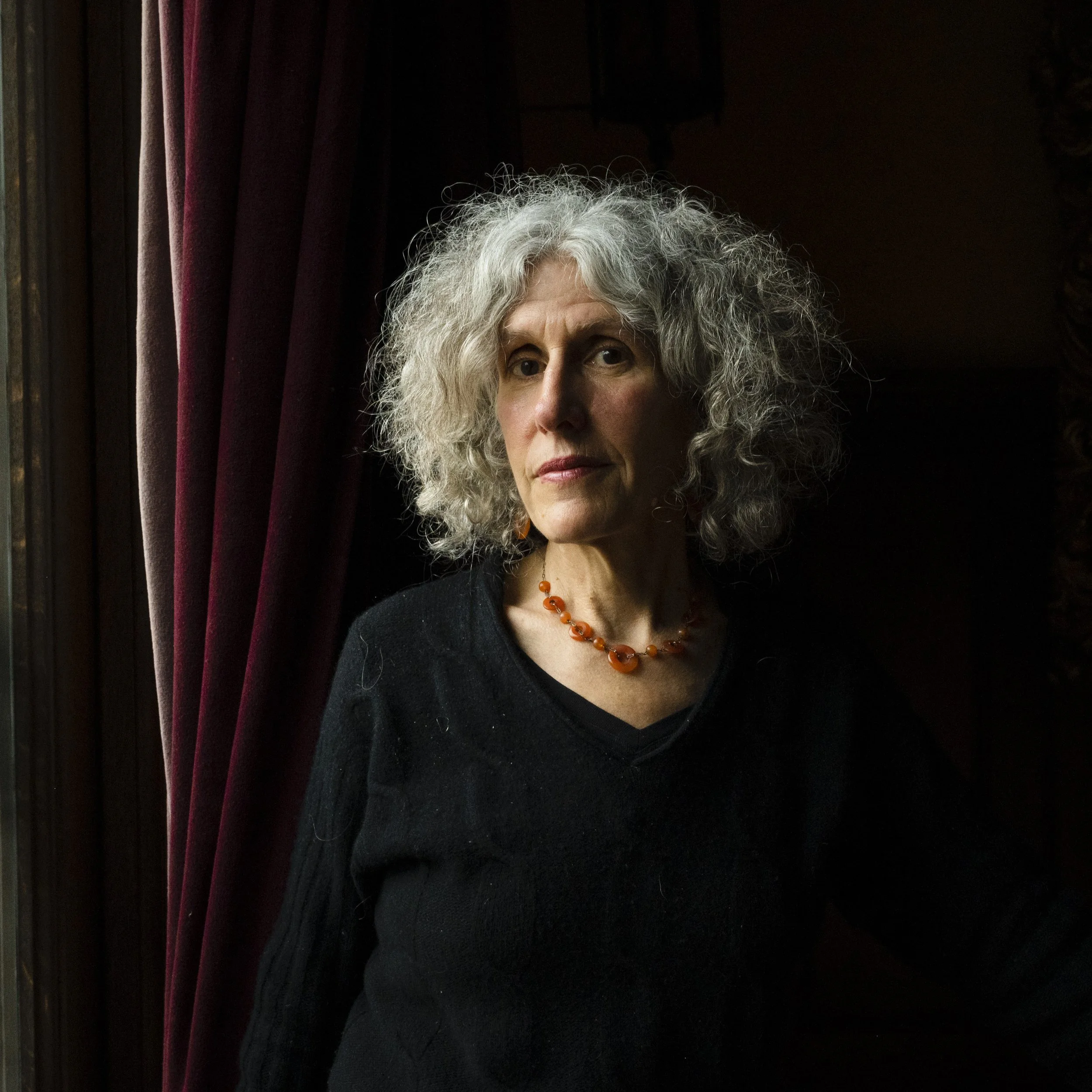 Portrait of an older white woman with curly gray hair, wearing a black sweater and an orange beaded necklace, standing near a dark curtain and looking towards the camera.