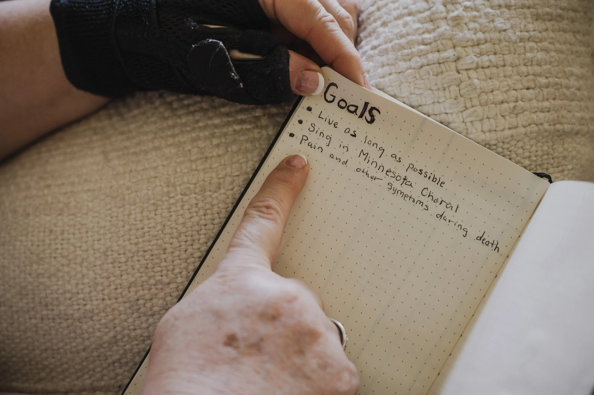 Close-up of a person's hand holding a small notebook page with handwritten notes about goals, including phrases like 'live as long as possible,' 'sing in ministry,' and 'pain and other symptoms during death,' on a textured beige surface.