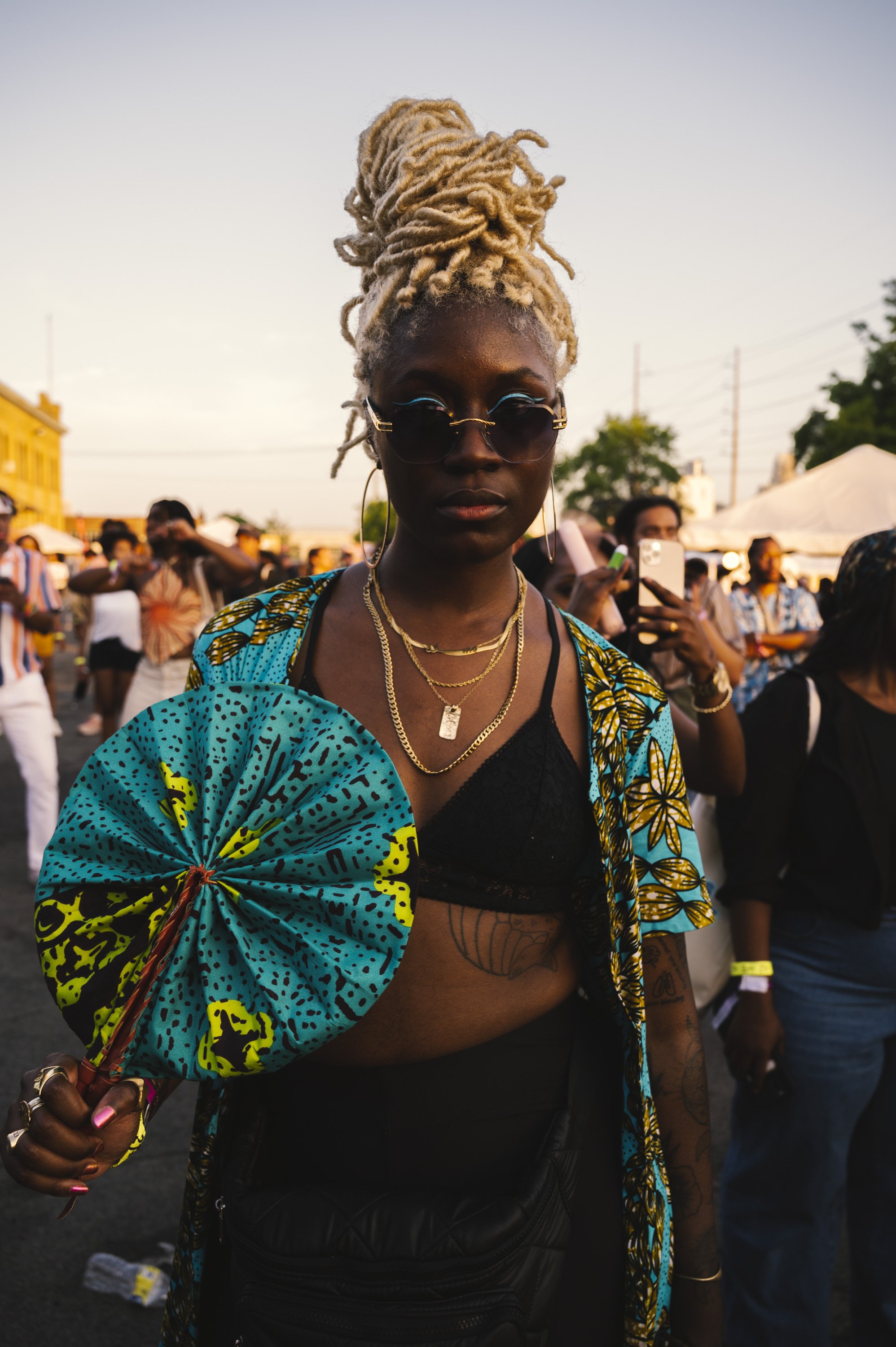 A woman with blonde dreadlocks styled in an updo, wearing sunglasses, multiple necklaces, a black crop top, and a colorful blue and yellow patterned shirt draped over her shoulders, standing outdoors at a crowded event during sunset.