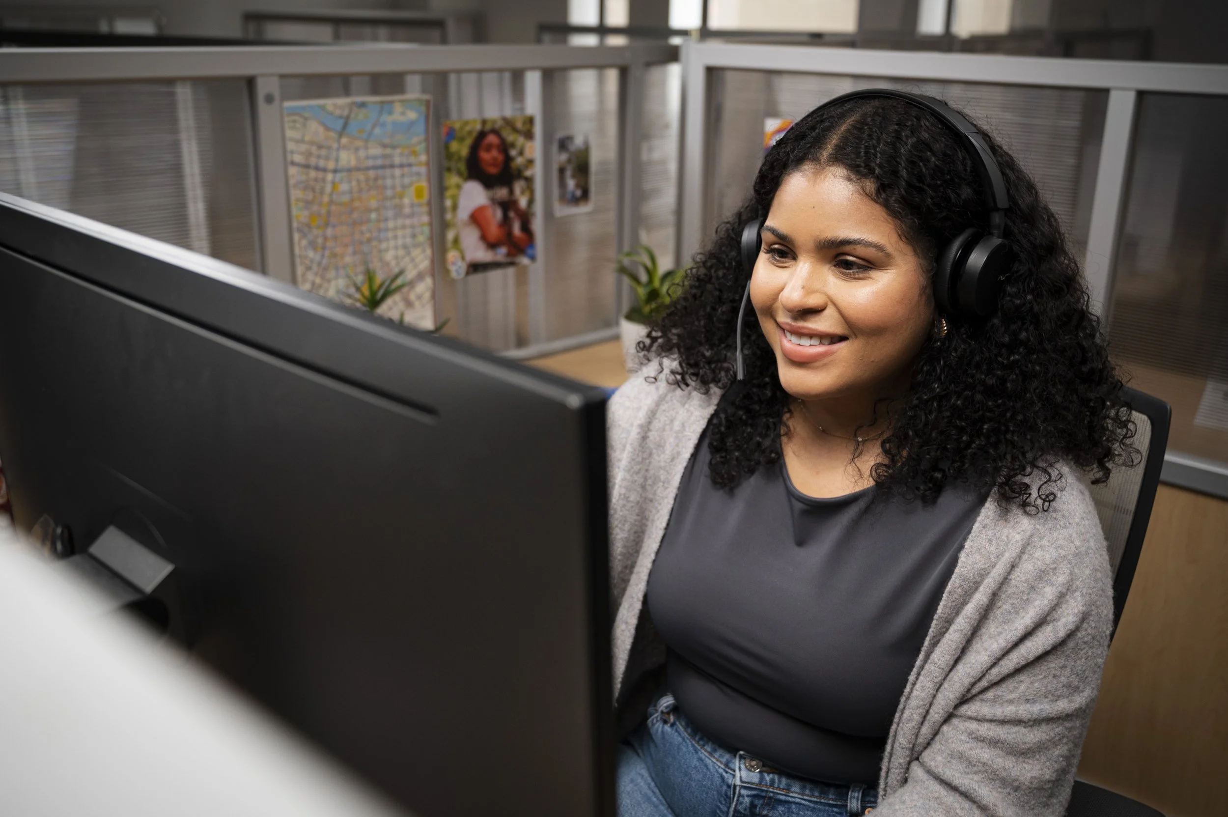 A racially ambiguous woman with curly black hair wearing a gray cardigan and black top, smiling while working at her computer in an office cubicle, with photographs and a plant on the background walls.
