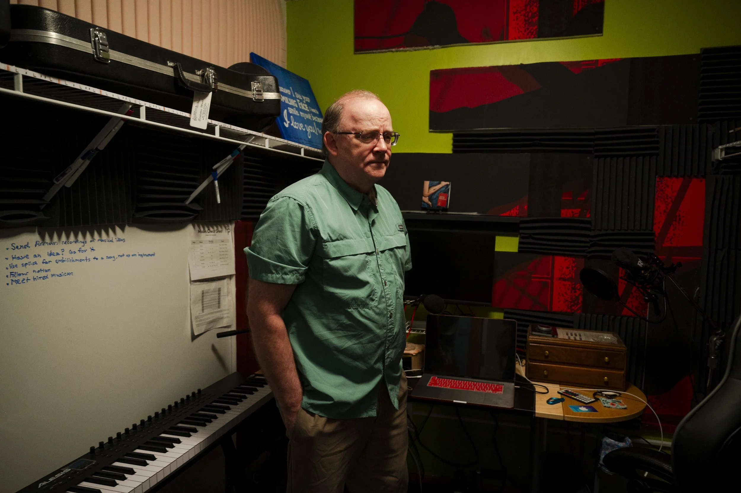 An older man with glasses wearing a green shirt stands in a music studio, surrounded by musical equipment including a keyboard, a microphone, and soundproofing foam panels on the walls.