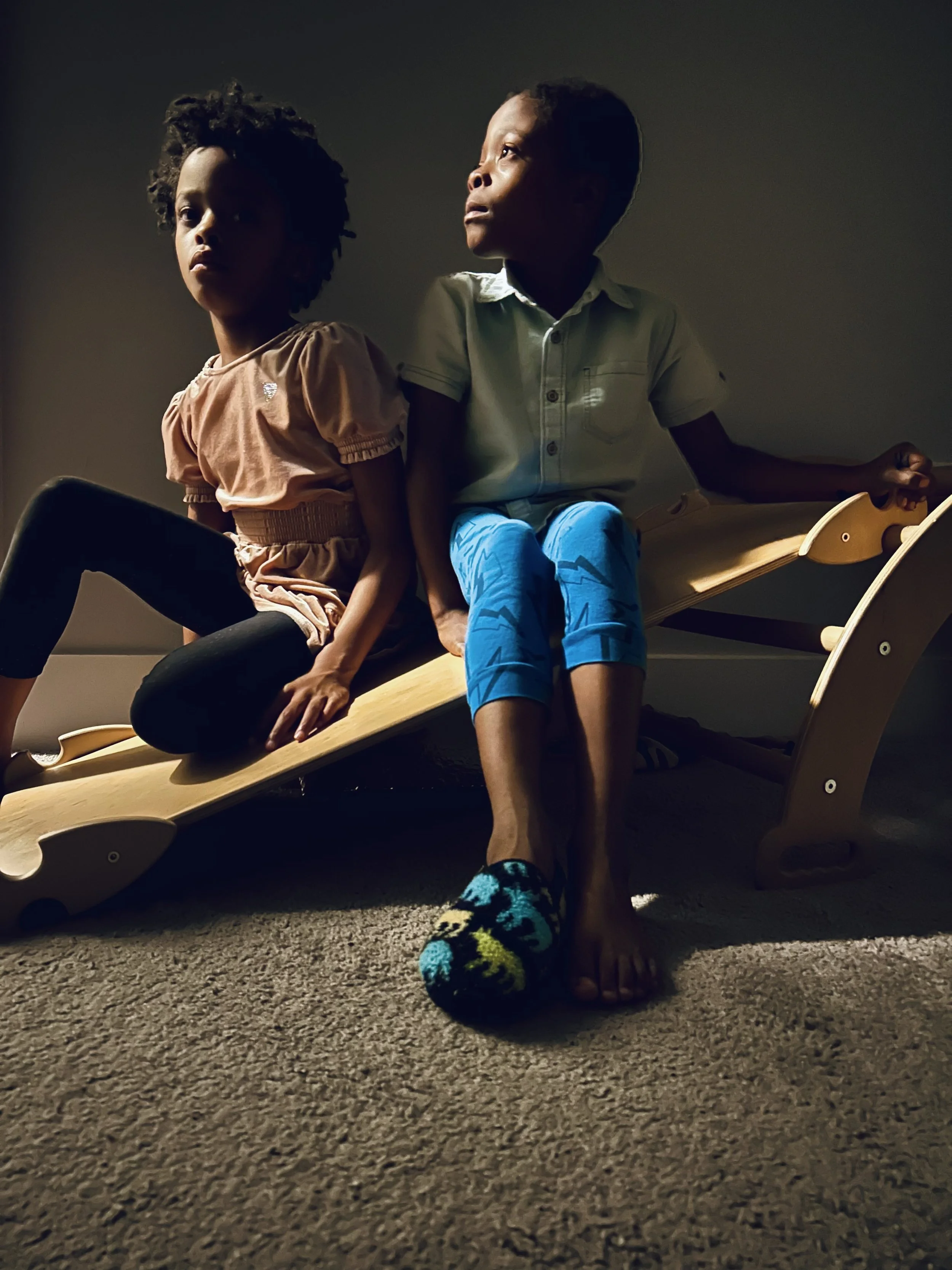 Two children sitting on a wooden swing in a dimly lit room, one wearing a pink dress and the other in a light shirt and blue pants, with the boy wearing colorful socks and barefoot.