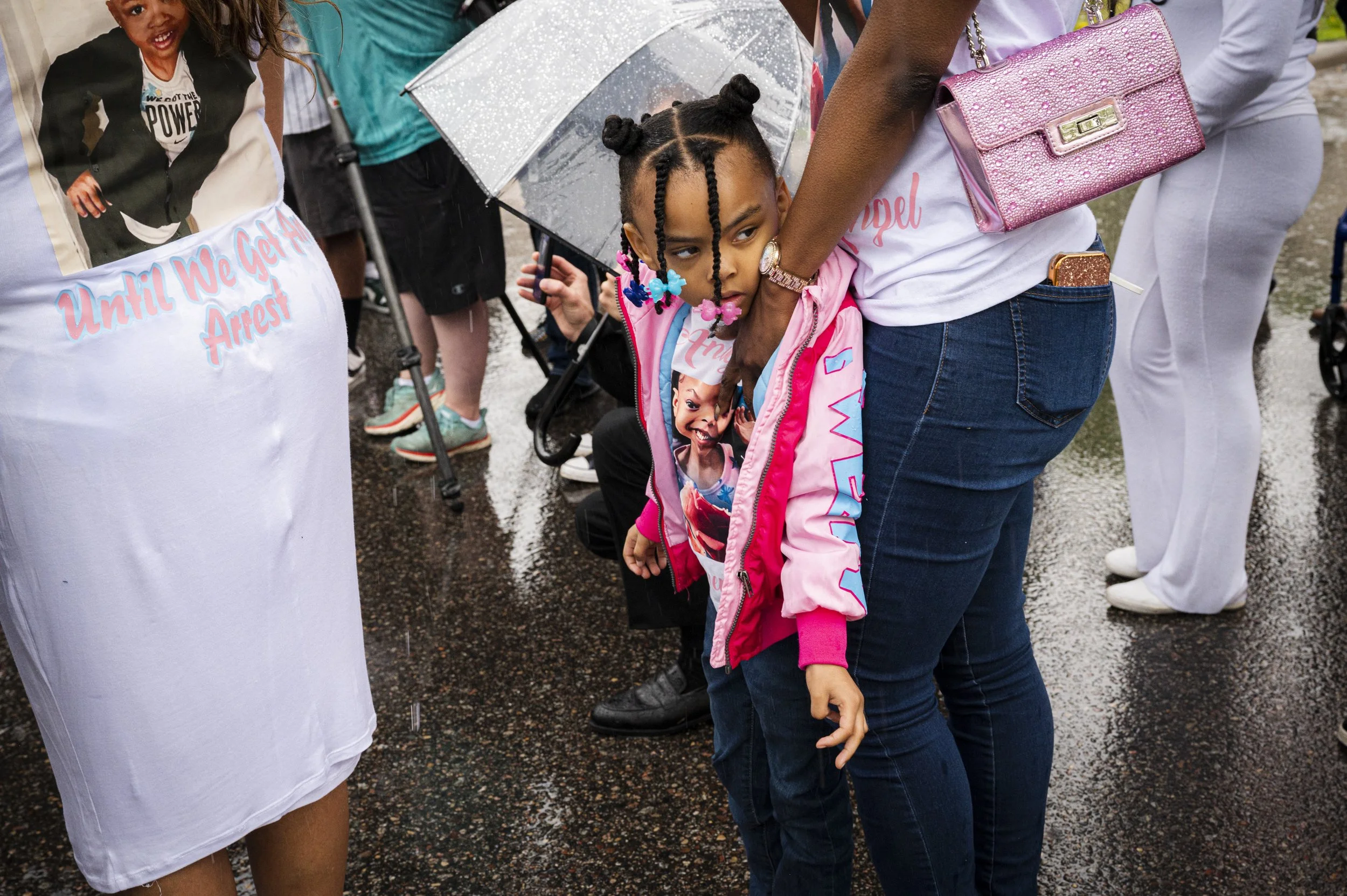 A young girl with braids, dressed in a pink jacket with a photo of a girl printed on it, holding onto an adult's waist, standing on a wet street during a rainy event. Several people around, some holding umbrellas, are visible in the background.