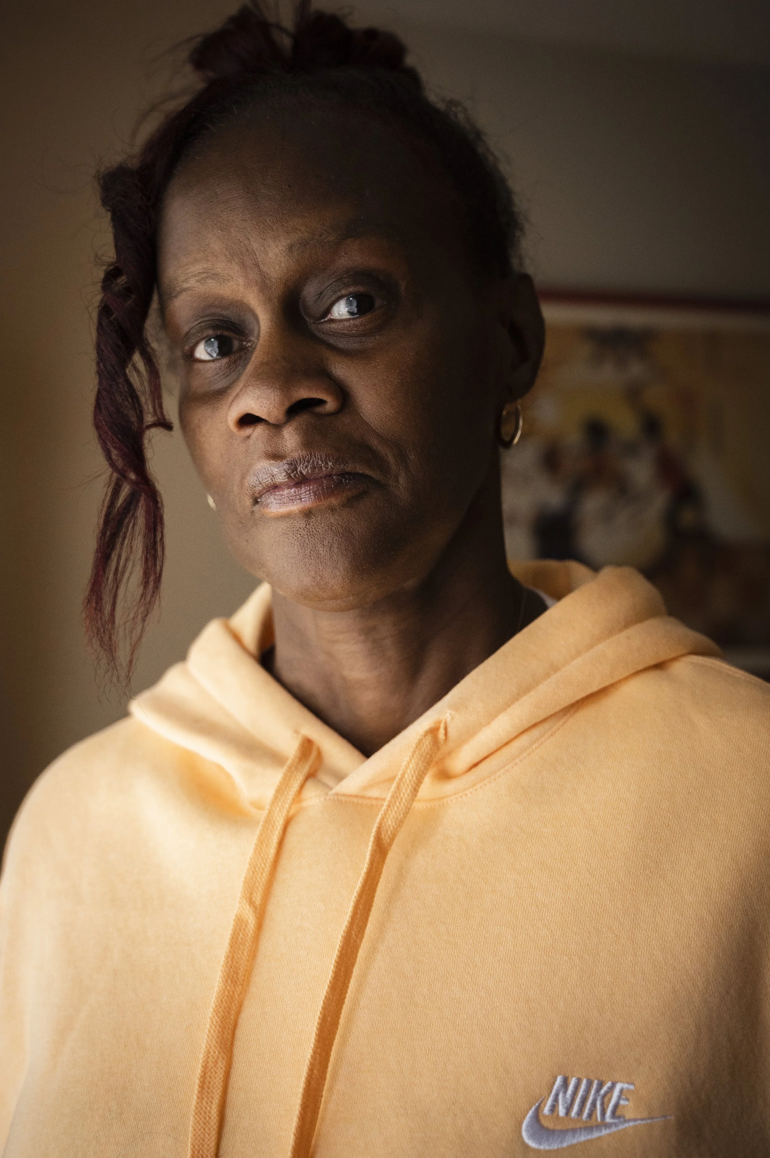 A woman wearing a peach Nike hoodie, looking at the camera with a serious expression, in a dimly lit room with a painting or poster in the background.
