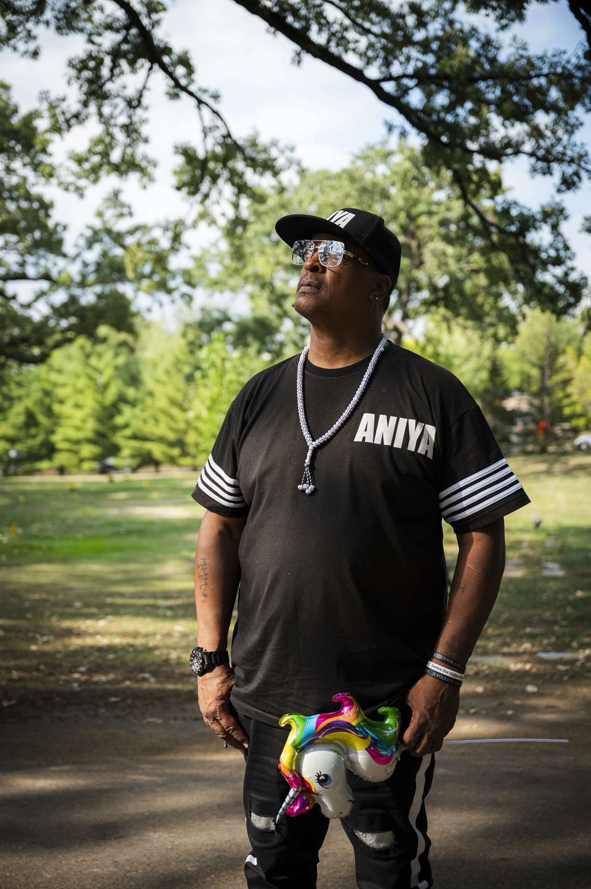 Man standing outdoors in a park, wearing a black ANIVIA t-shirt, black cap, sunglasses, and jewelry, holding a colorful unicorn-shaped balloon.
