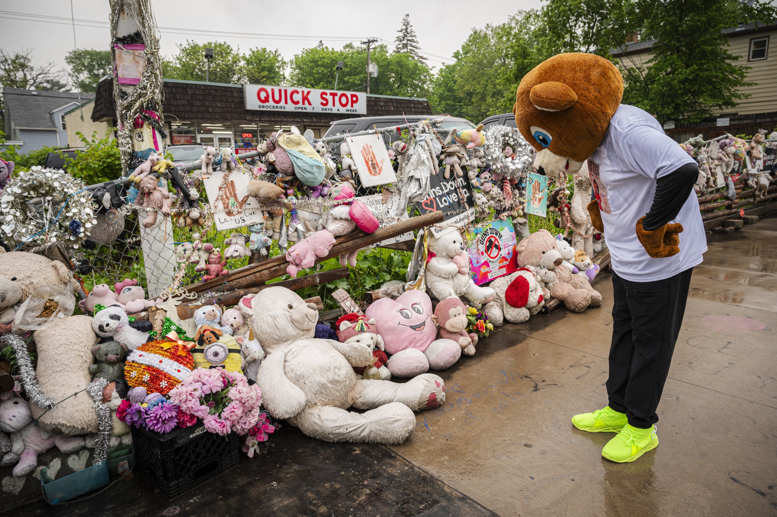 Person dressed in a bear mascot costume looking at a memorial of stuffed animals and flowers outside store.