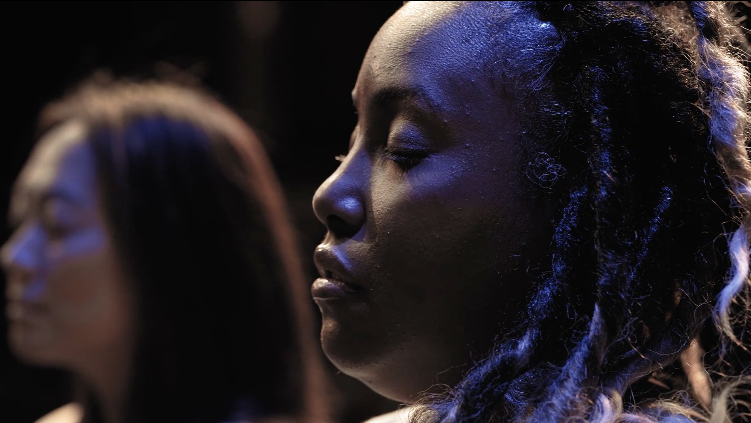 Close-up of a woman with her eyes closed, facing sideways with emphasized dark skin and braided hair, lit with bluish lighting.