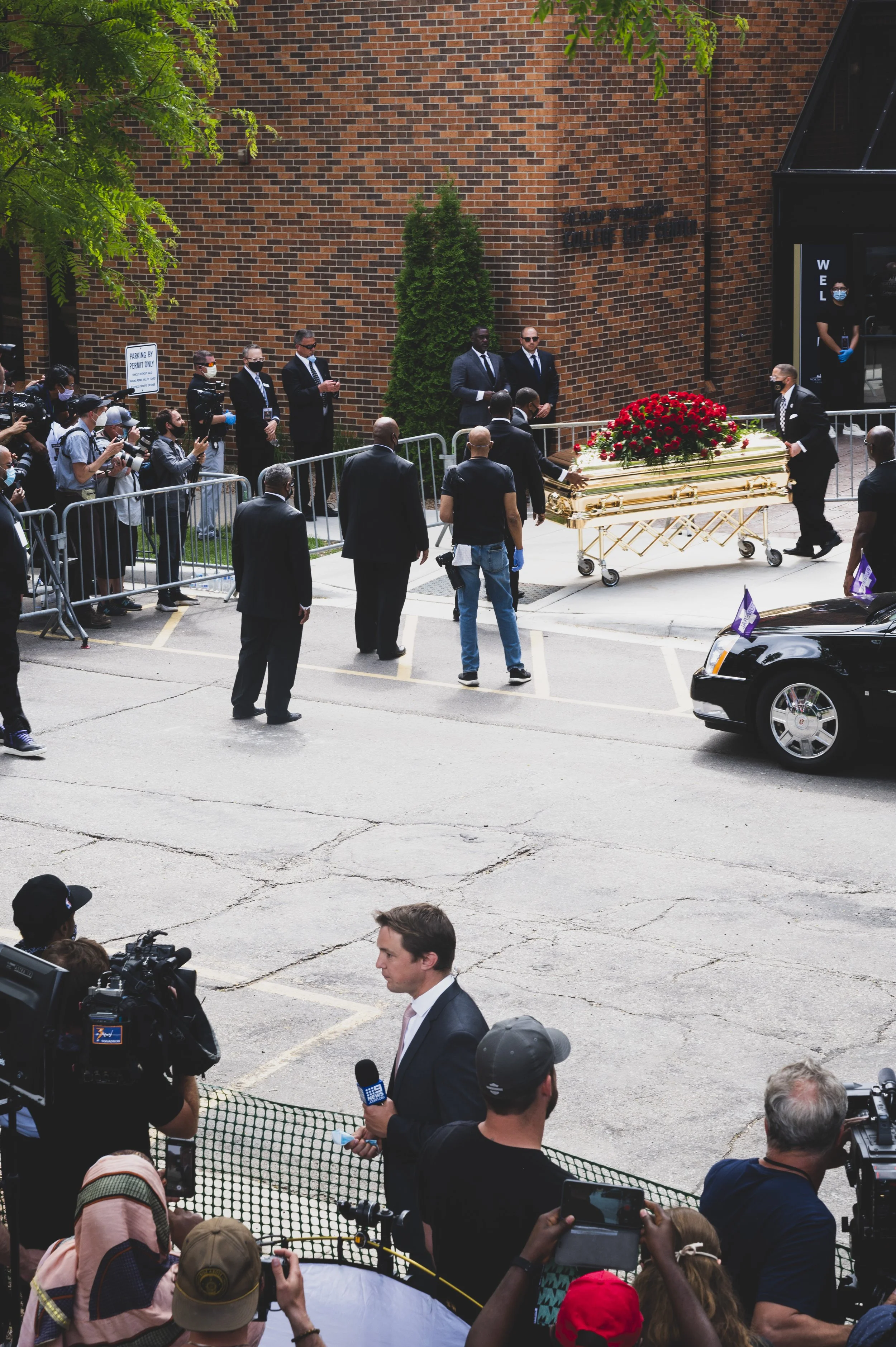 A funeral procession with a golden casket adorned with red roses being carried by pallbearers. People are gathered around, some wearing masks, with the scene taking place outside a brick building. A man in a suit is speaking into a microphone, and ph