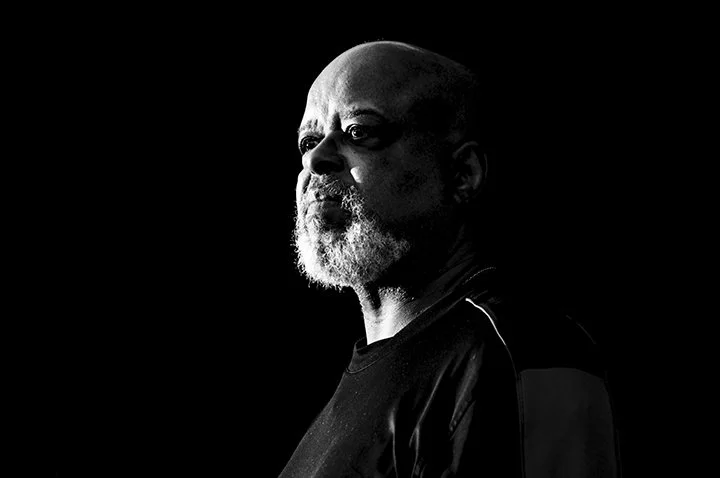Black and white portrait of a bald man with a gray beard, looking to the side, with a serious expression, shirt with shiny fabric, and a dark background.
