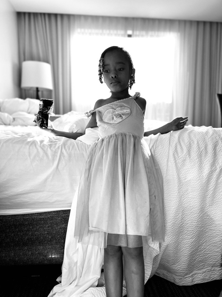 A young girl in a dress standing in a hotel room next to a bed with a chandelier in the background
