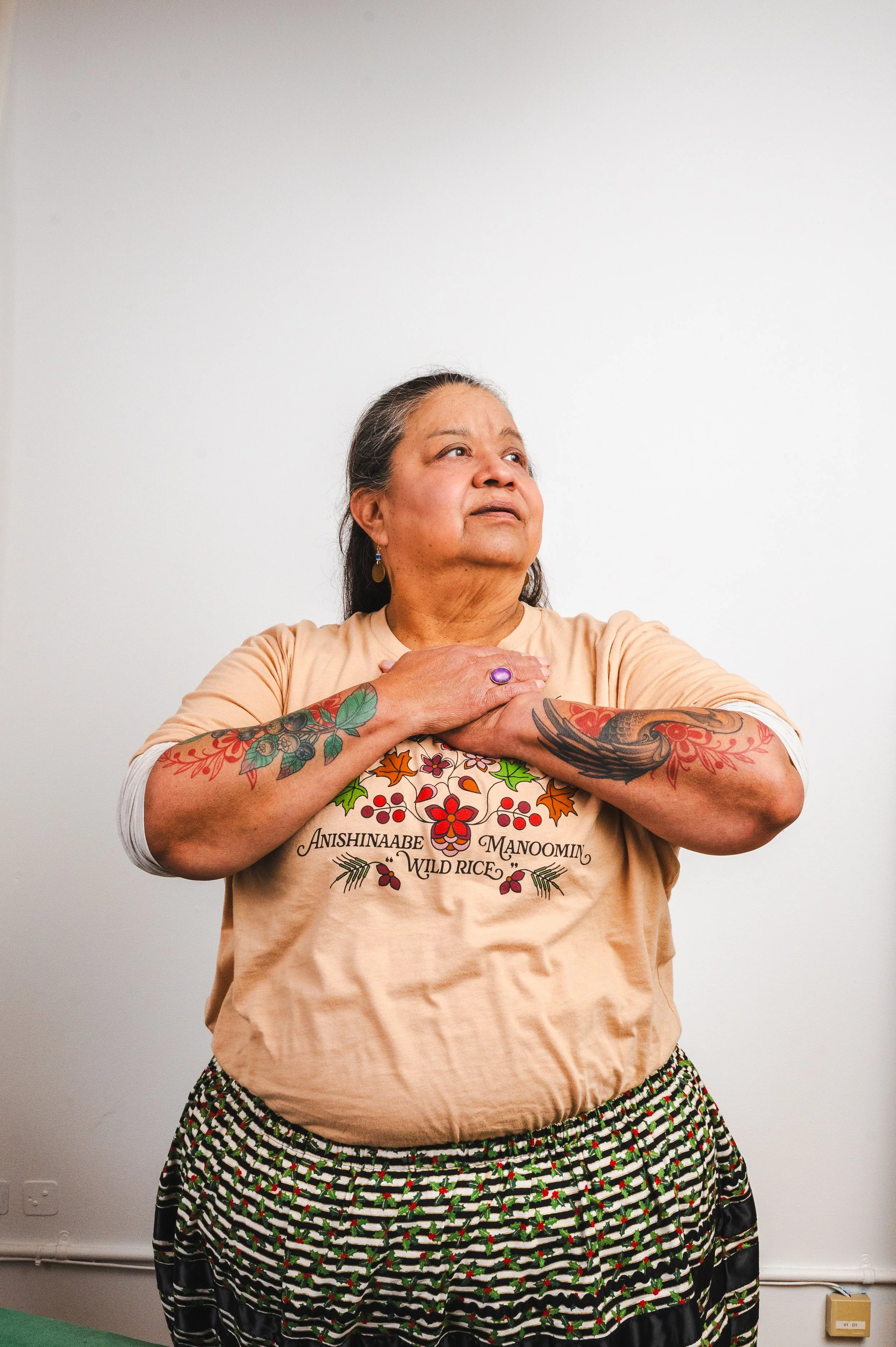 A woman with long dark hair, tattoos on her arms, wearing a beige T-shirt and patterned skirt, stands against a plain white wall, holding her hands over her heart and looking upward.