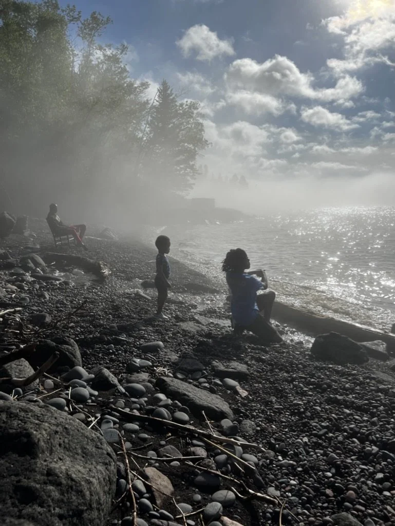 A foggy lakeside scene with a teenager, a boy, and a man sitting on a chair, involving rocky terrain, water, trees, and a partly cloudy sky.