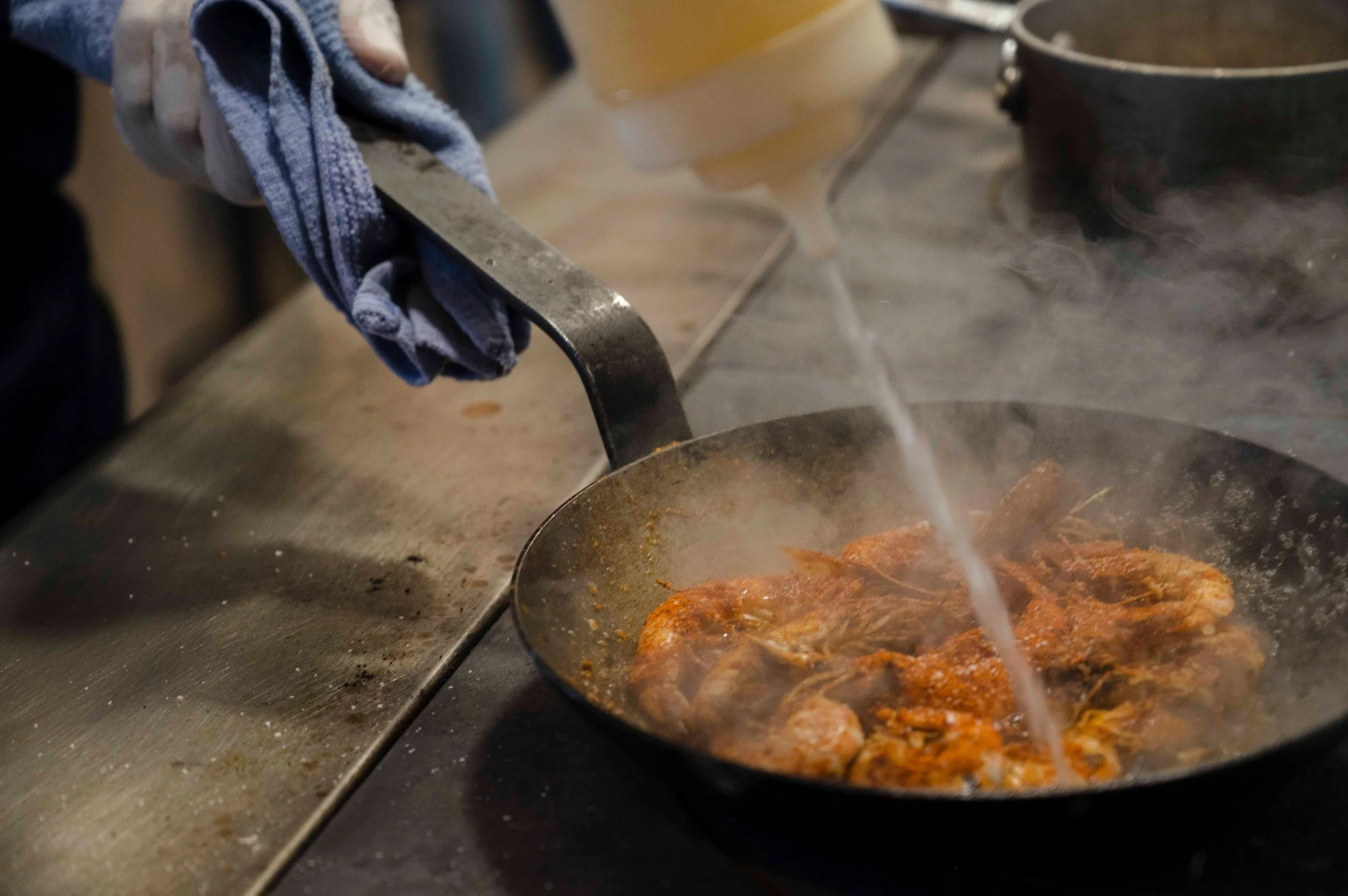 A person cooking shrimp in a skillet with a splash of water being poured in, with steam rising from the food.