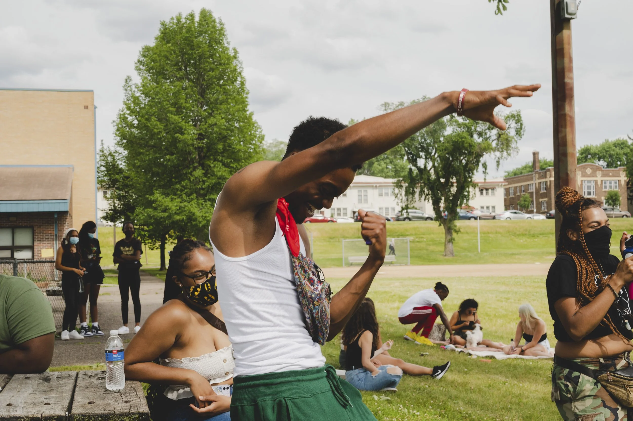 A group of diverse people at an outdoor gathering on a grassy field. In the foreground, a person wearing a white tank top, green pants, and a red scarf is dancing or celebrating with their arm raised. Other individuals are sitting or standing, some w
