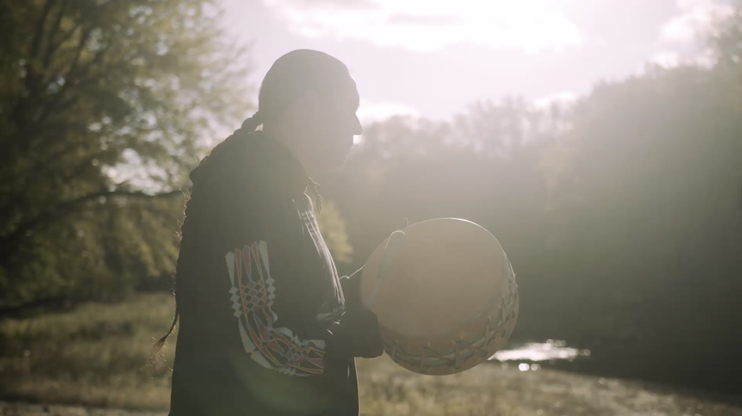 A Native American man holding his tribal drum in front of the Mississippi River at sunset, with trees in the background.