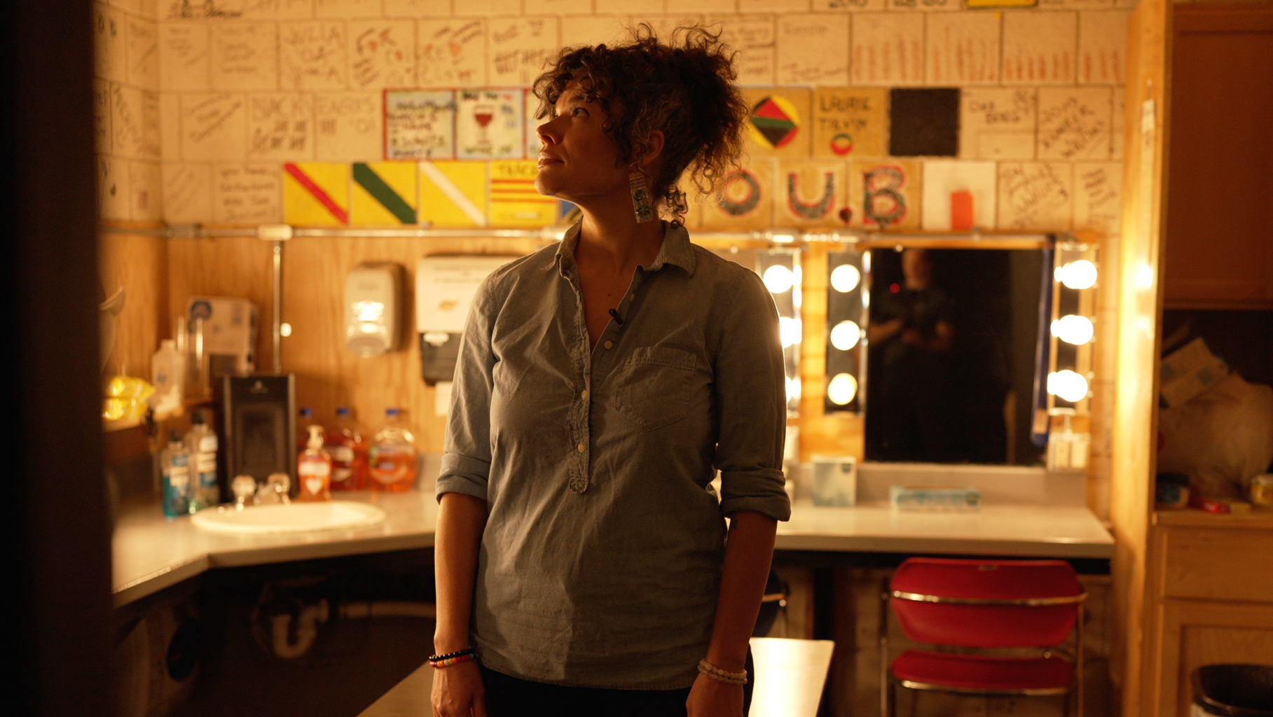 A woman standing in a dressing room with a mirror and bright lights, surrounded by signed posters and memorabilia on the walls.