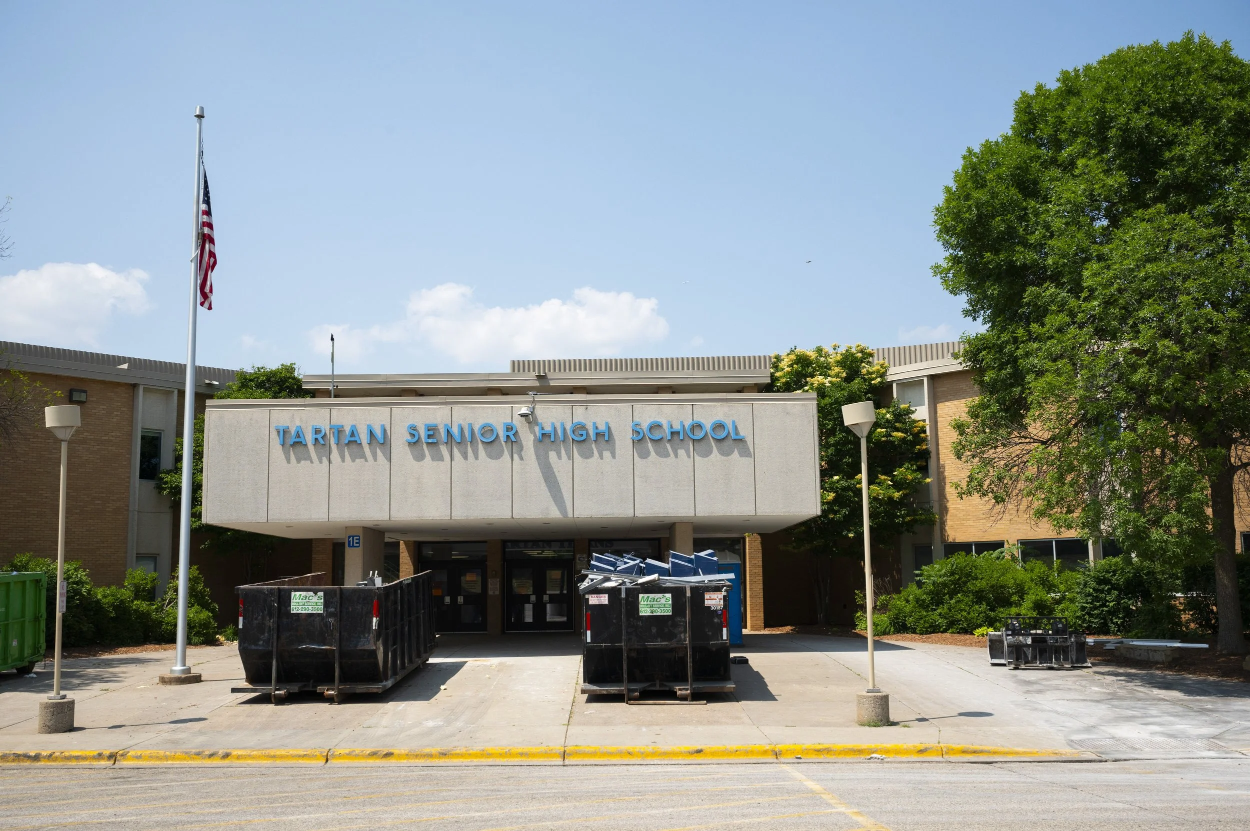 Front view of Tartan Senior High School building with blue sky, trees, and flagpole with American flag. There are containers and dumpsters in the foreground and recycling bins on either side of the entrance.