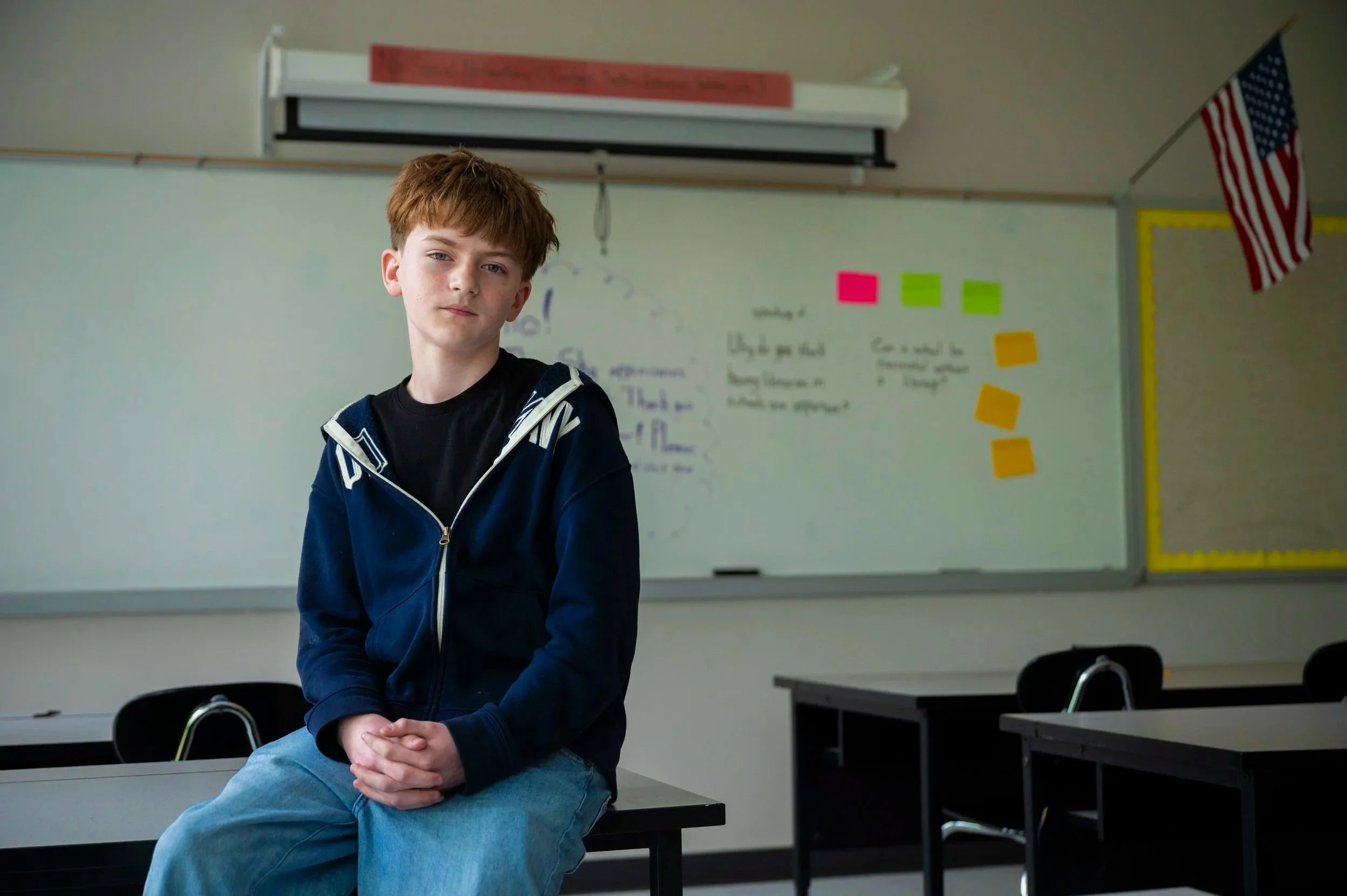 A young boy with reddish-brown hair, wearing a black shirt and a blue zip-up hoodie, is sitting on a desk in a classroom, facing the camera with a neutral expression. The classroom has white walls, a whiteboard with colorful sticky notes, and an American flag hanging on the side.