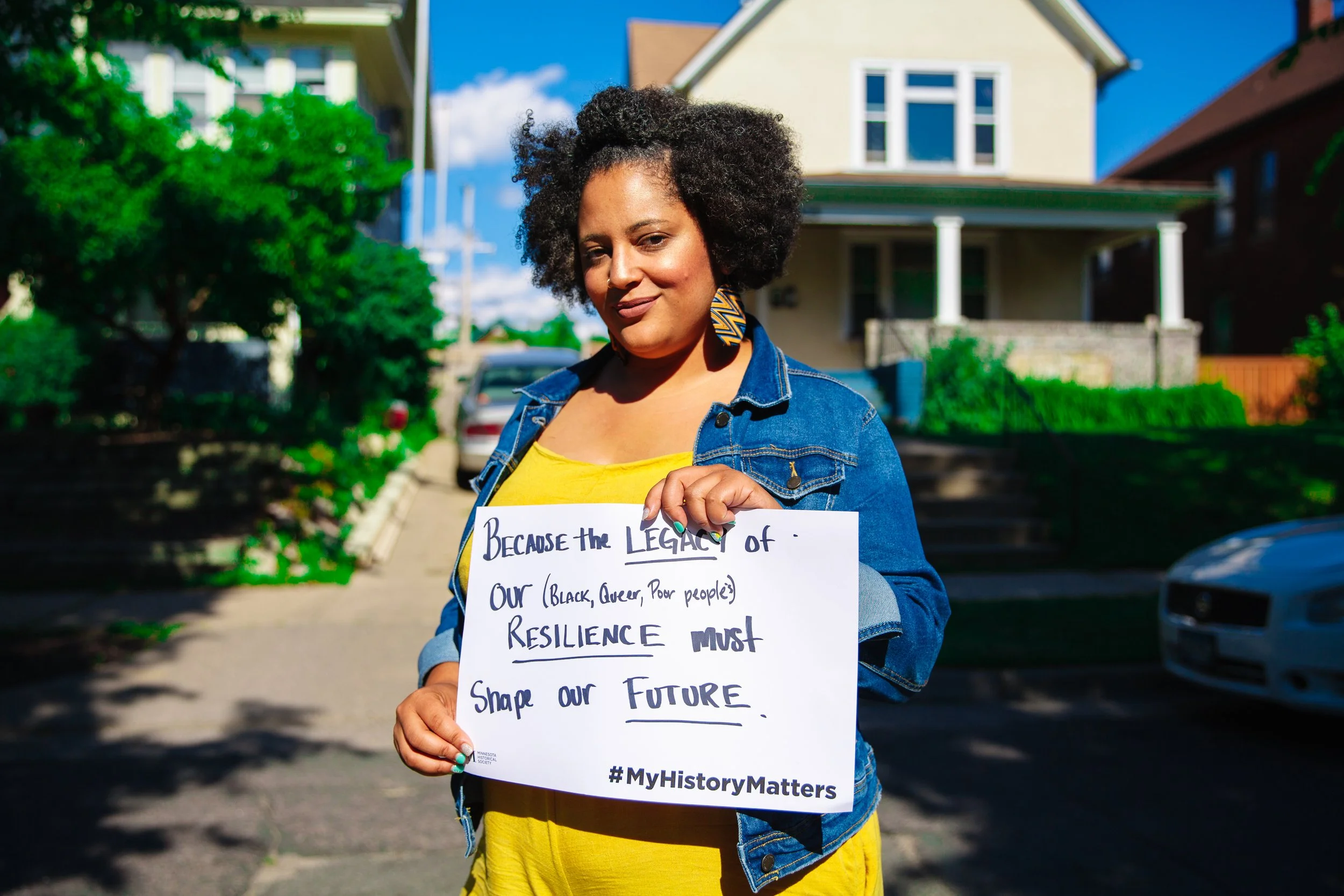 A woman with natural hair wearing a yellow shirt and denim jacket holding a sign that reads 'Because the legacy of our (Black, queer, poor people's) resilience must shape our future' standing outdoors on a sunny day.