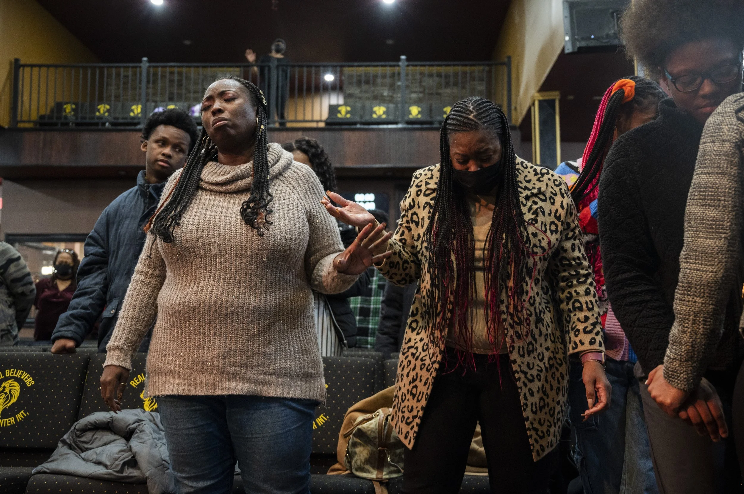 A diverse group of people is praying or reflecting, with eyes closed, in a church or similar setting. They are standing with their heads bowed and hands raised or clasped together.