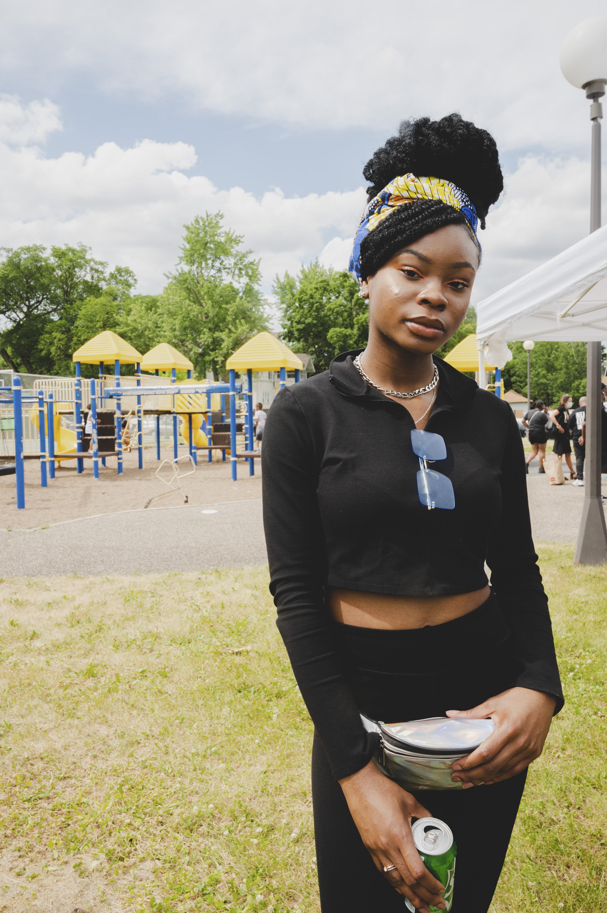 A young woman with a colorful head wrap, gold necklaces, and a black crop top standing outdoors at a park with playground equipment, trees, and other people in the background, holding a can and a holographic purse.
