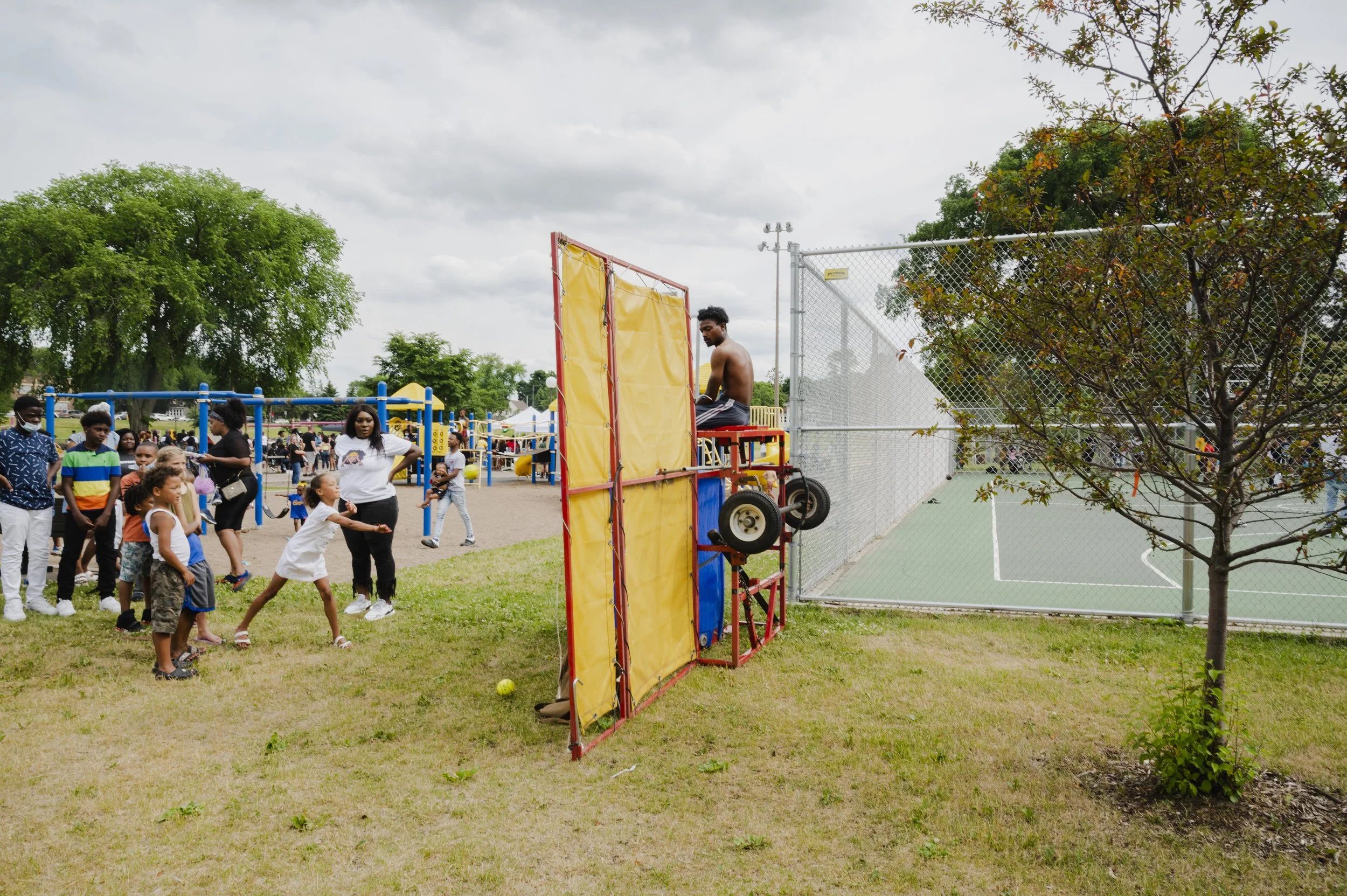 A public park with a large group of children and adults gathered around a mobile tennis ball shooter. A shirtless man is sitting on a red platform, operating the machine. There are trees, a chain-link fence surrounding a tennis court, and a cloudy sk