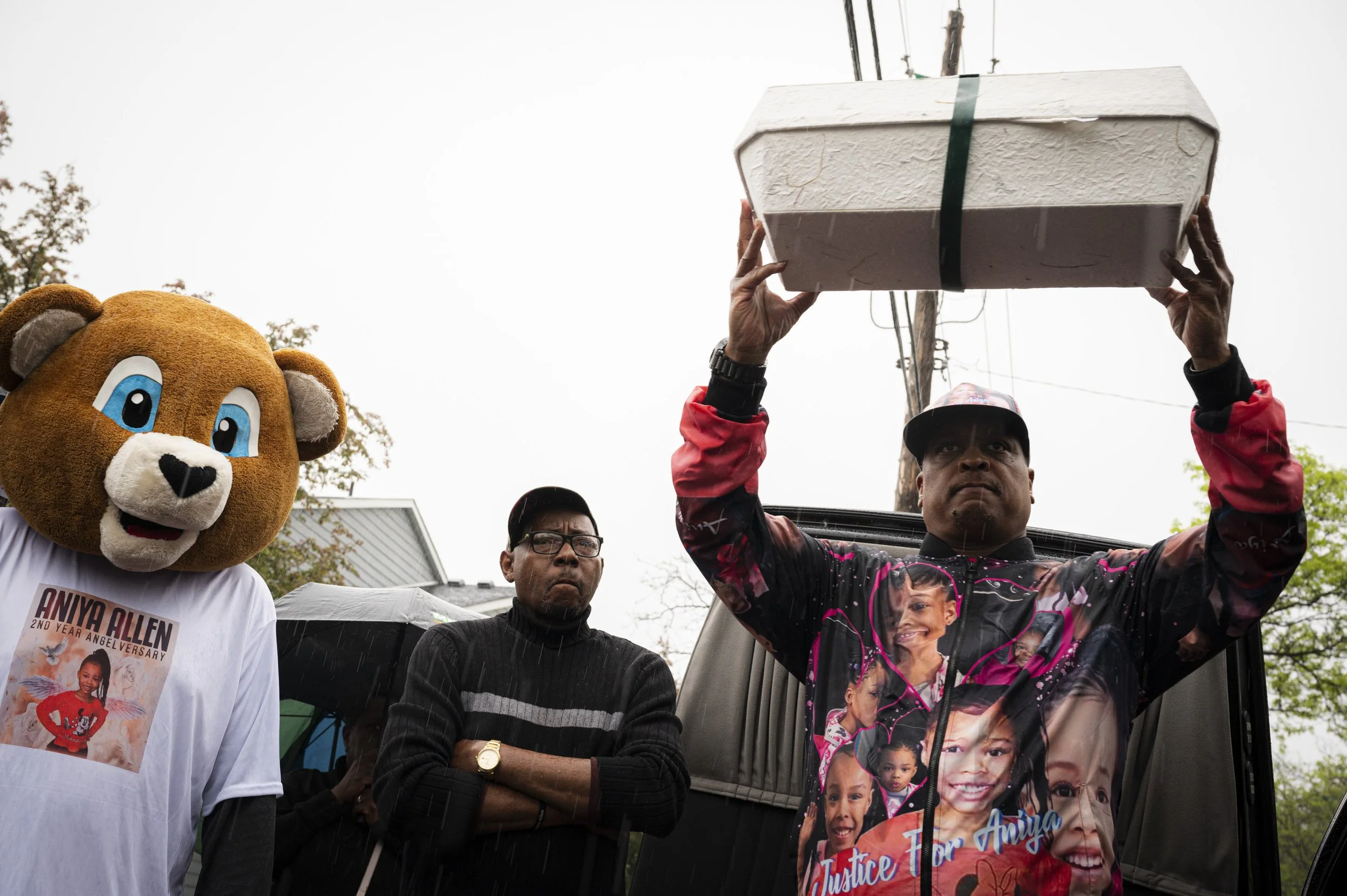 A person wearing a colorful jacket holds a large box above their head, standing next to two men, one of whom wears a black turtleneck, and a person in a bear mascot costume wearing a white T-shirt with a photo of a young girl and text that reads 'Any