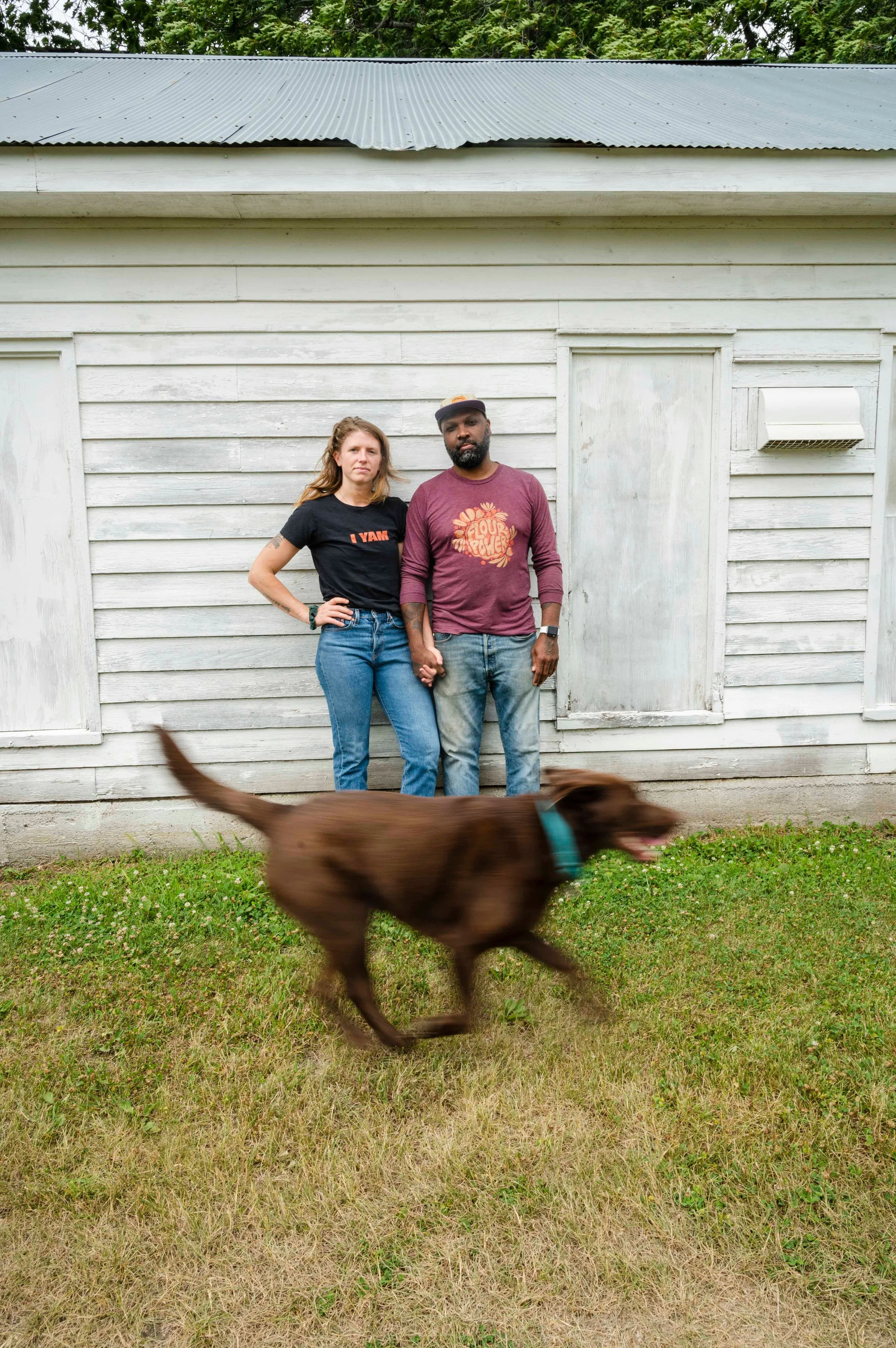 A man and woman standing hand-in-hand in front of a weathered white wooden building, with a brown dog running across the grass in front of them.