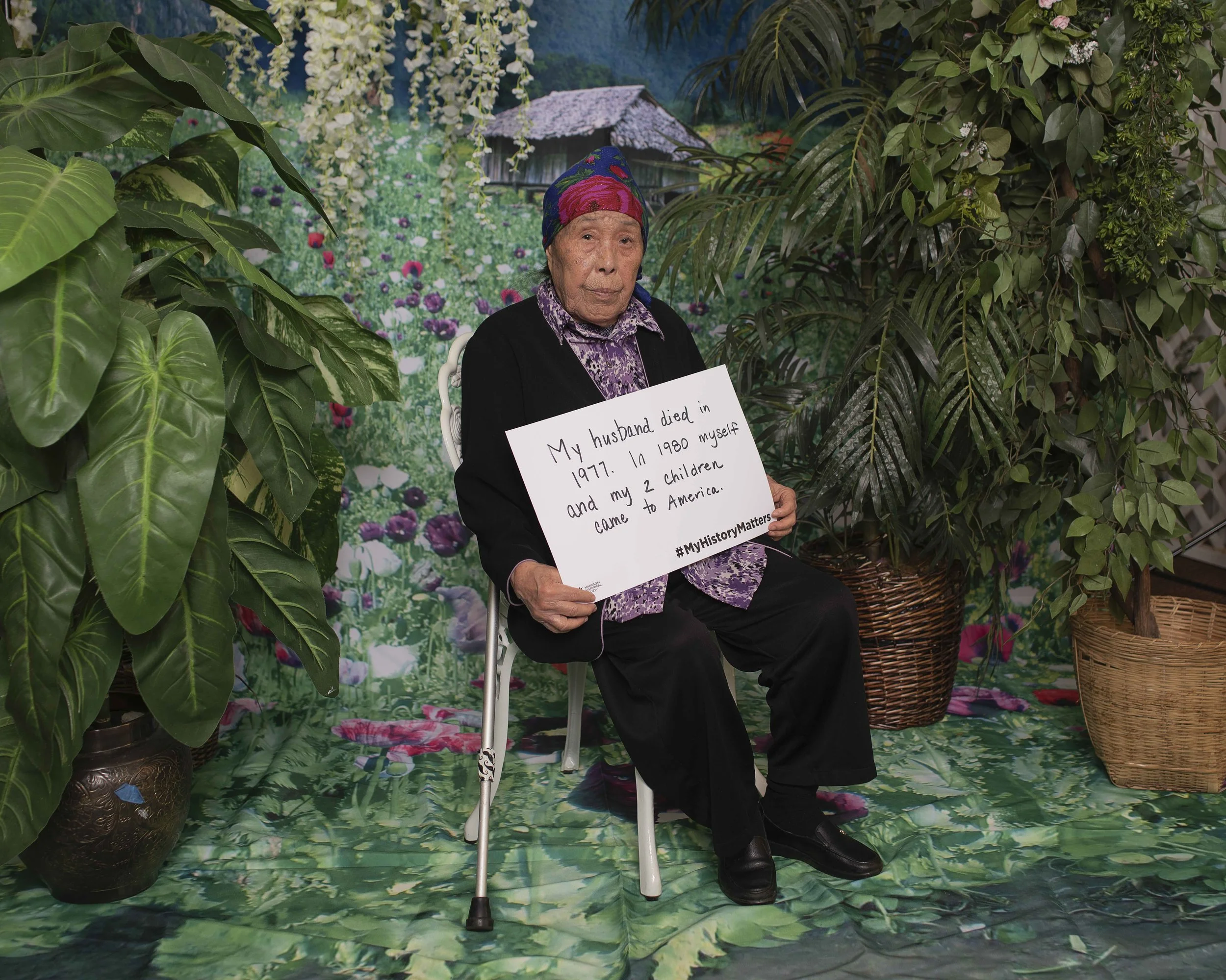 An elderly woman sitting on a white chair in front of a colorful garden backdrop, holding a sign that reads, 'My husband died in 1977. In 1980 myself and my 2 children came to America. #MyHistoryMatters'. The setting includes large green plants and a