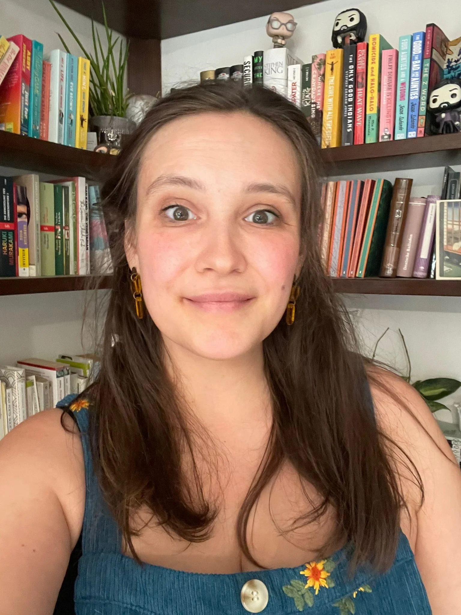Junge Frau mit braunen, glatten Haaren trägt eine blaue Bluse mit Blumenmuster, befindet sich vor einem Bücherregal mit bunten Büchern und Figuren.