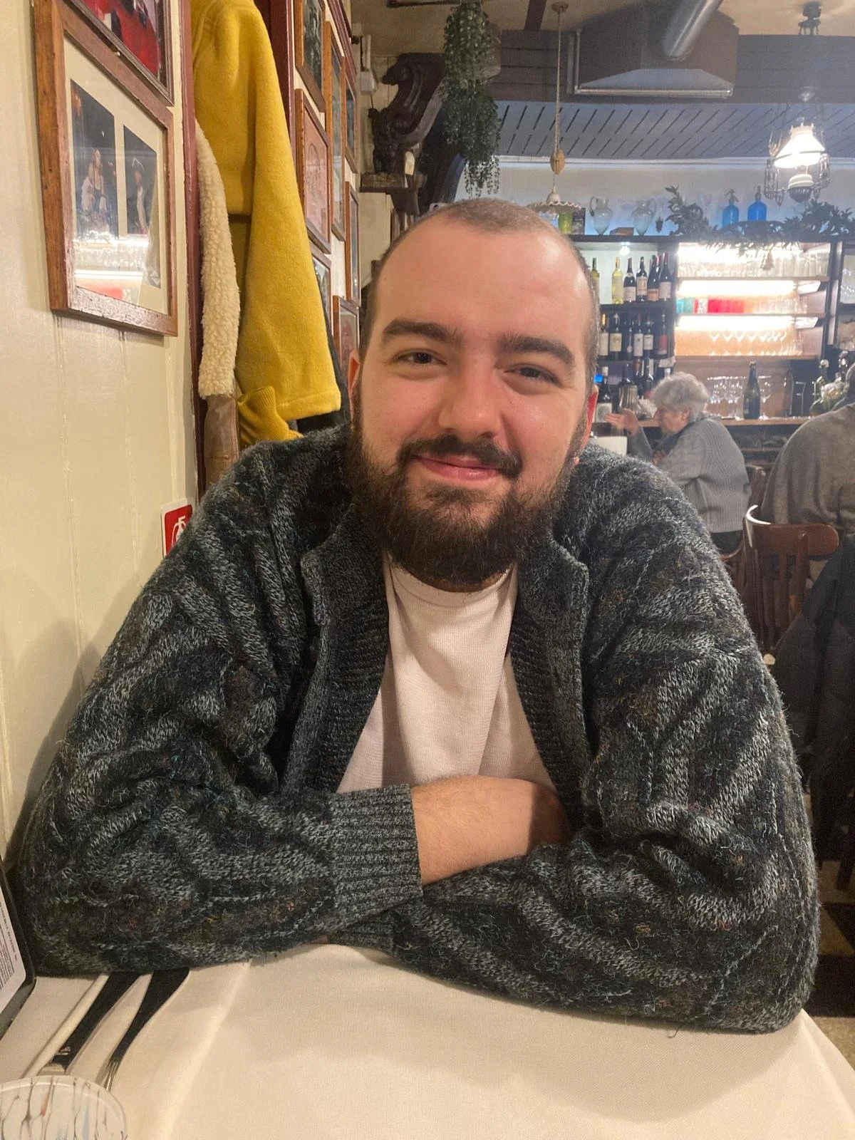 Ein Mann mit dunklem Haar und Bart sitzt an einem Restauranttisch, trägt einen grauen Pullover und lächelt. Im Hintergrund sind andere Gäste, Weinflaschen und Dekorationen sichtbar.