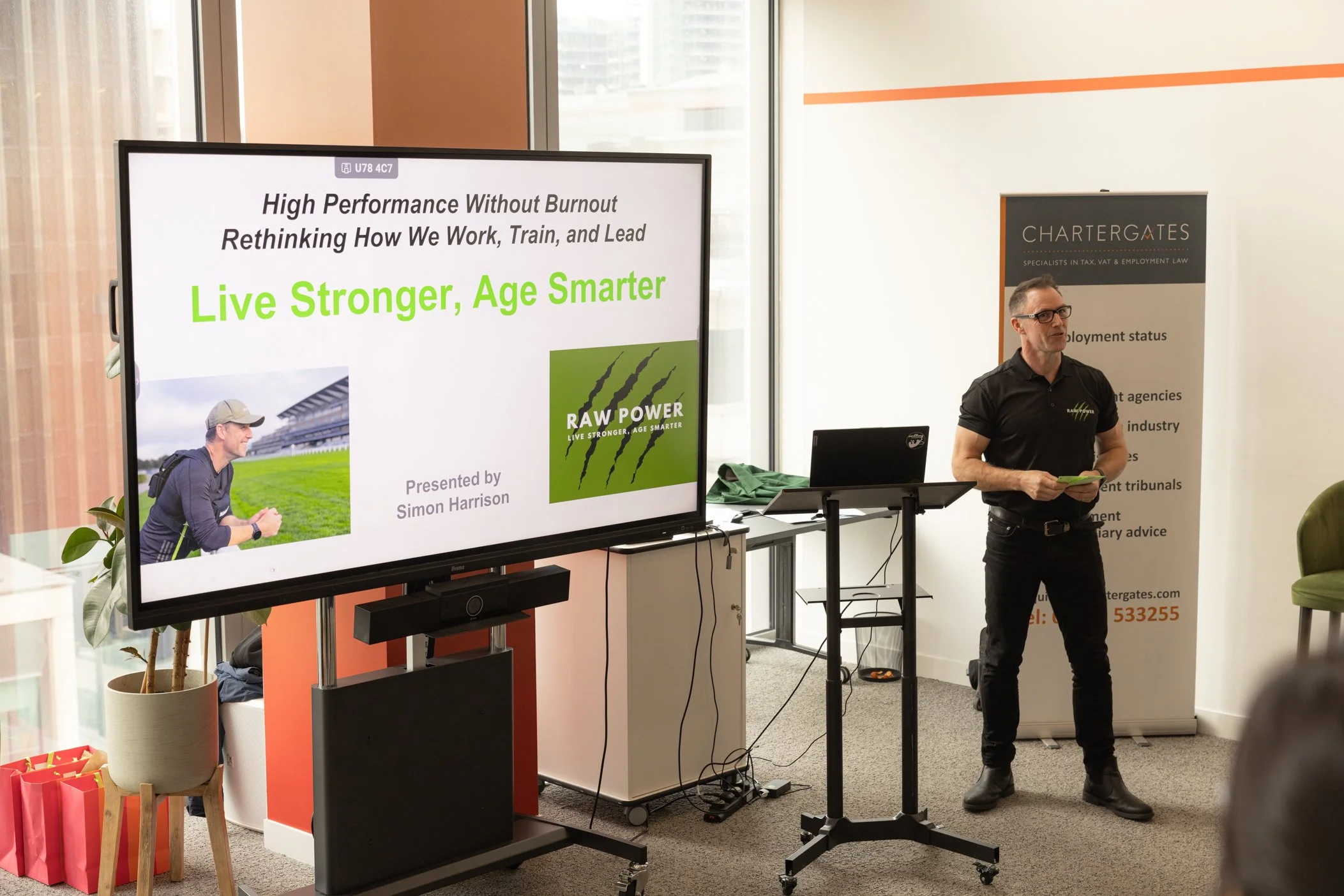 A man in a black shirt and glasses standing in front of a banner and a large screen with a presentation slide. The slide reads 'High Performance Without Burnout, Rethinking How We Work, Train, and Lead, Live Stronger, Age Smarter,' with an image of a person outdoors and a green logo for RAW POWER.