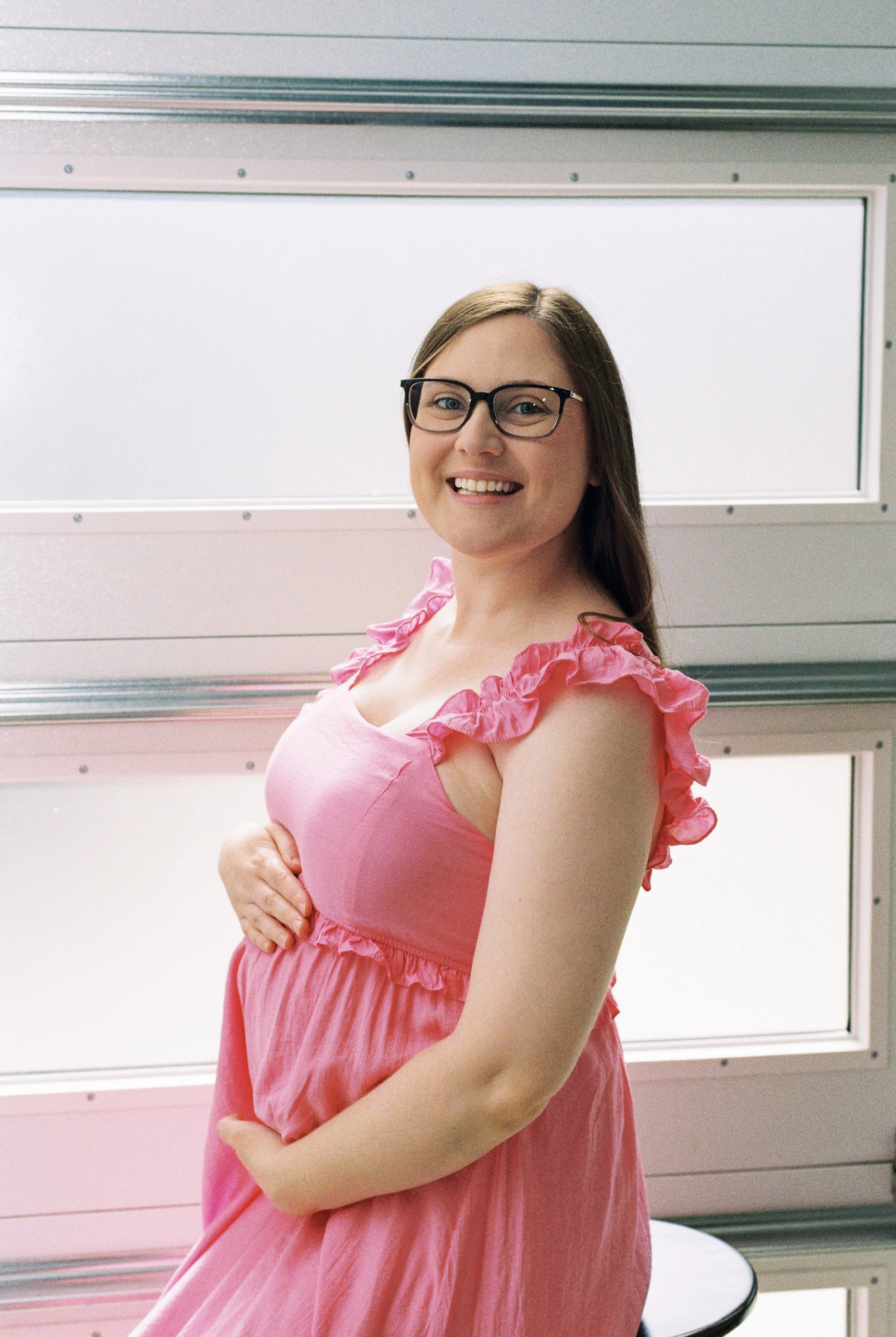 A woman with glasses wearing a pink dress with ruffled sleeves, smiling with one hand on her belly, standing in front of a metallic and white panel background.