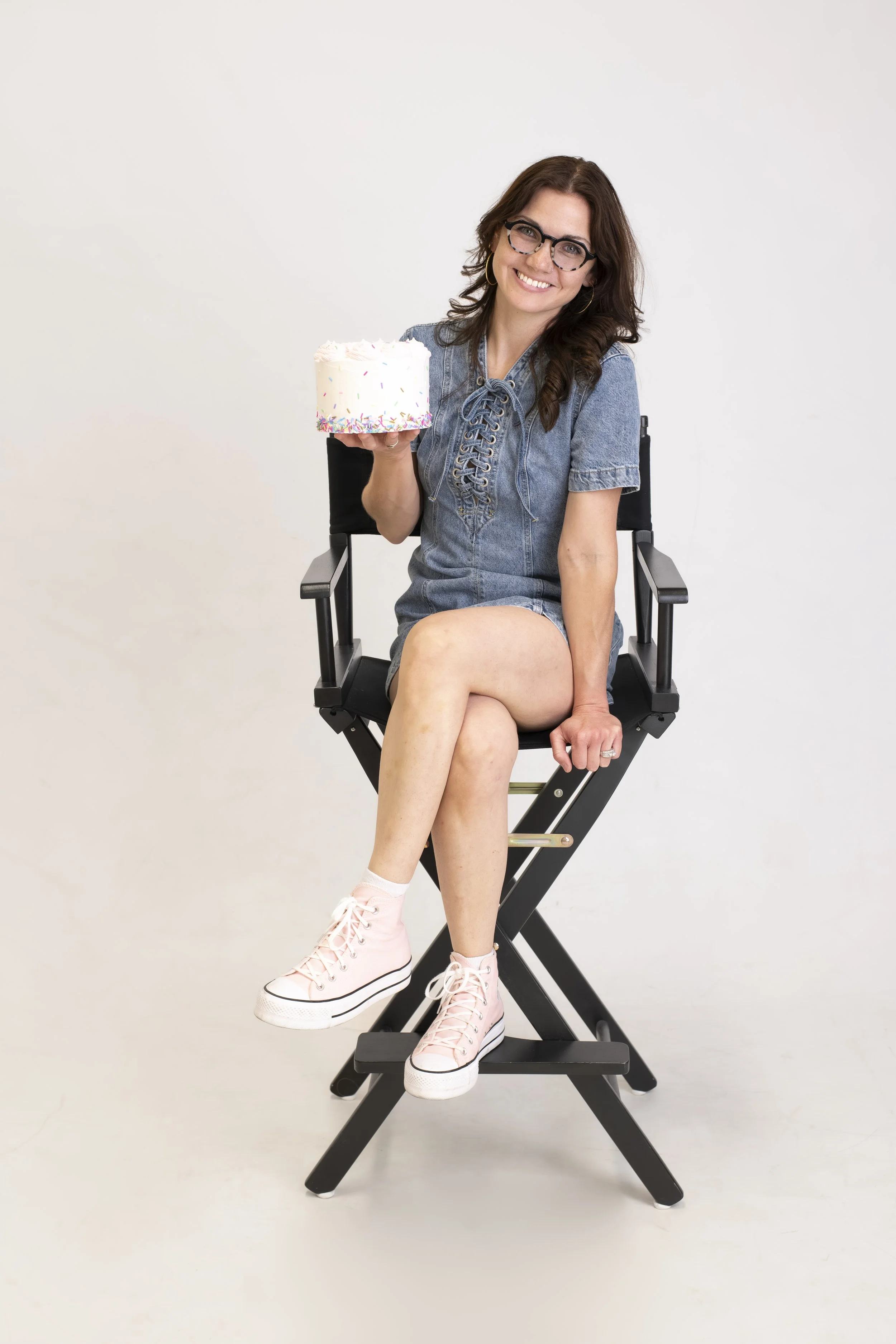 Woman sitting on a director's chair, holding a birthday cake with sprinkles, smiling, wearing glasses, pink high-top sneakers, denim dress, in a plain white background.