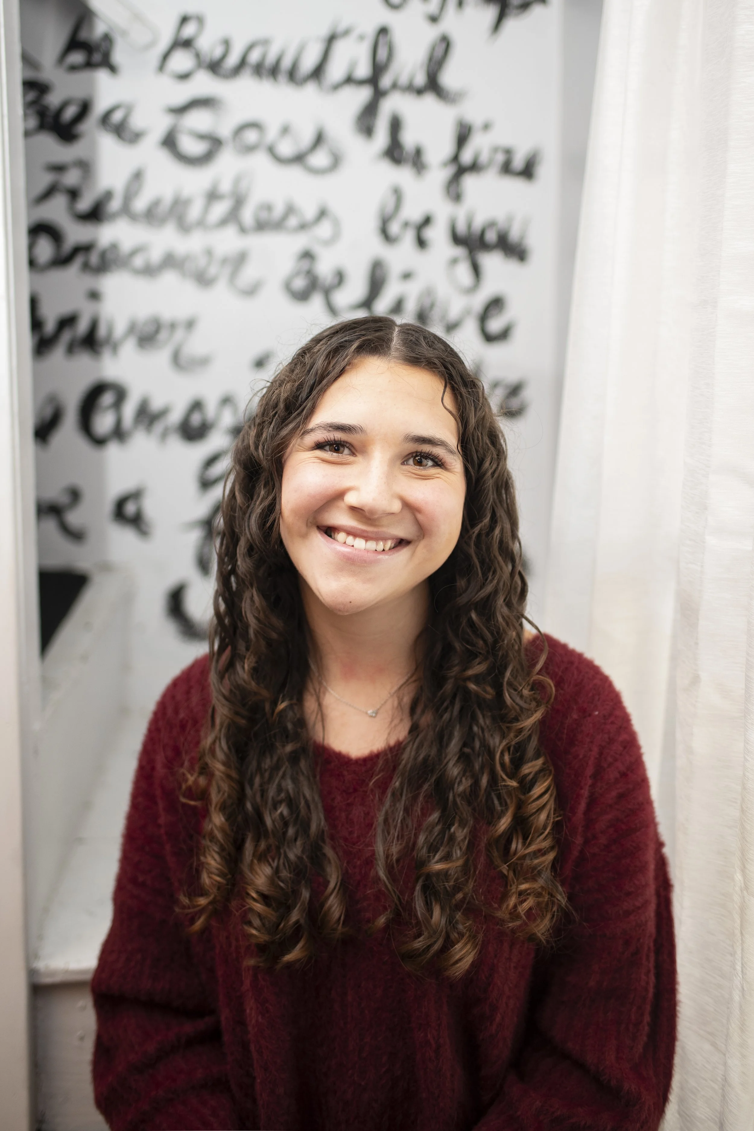 A young woman with long, curly brown hair and a big smile, wearing a maroon sweater, sitting in front of a mirror in a room with decorative wall art and white curtains.